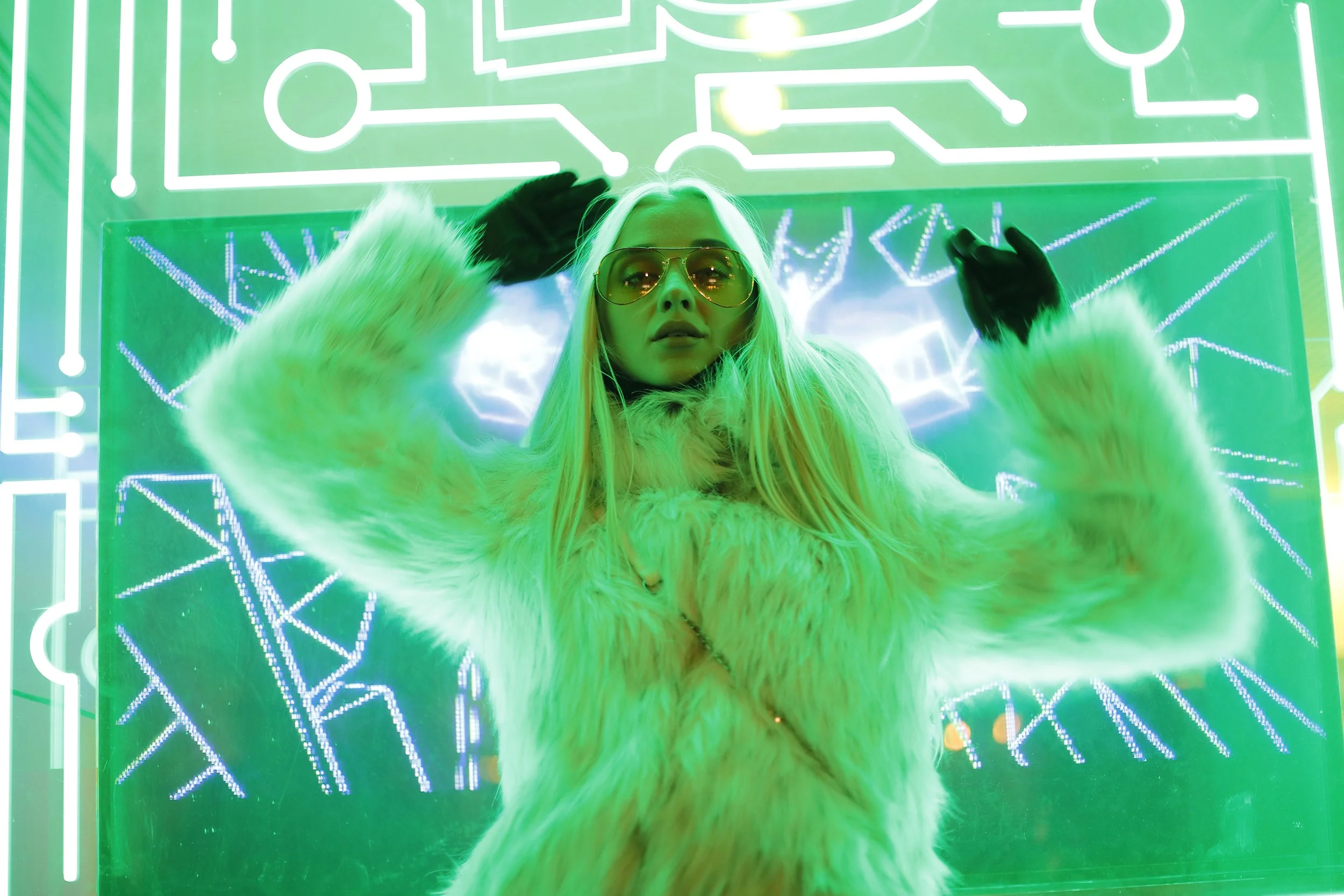 A woman with long blonde hair and sunglasses, wearing a green fur coat and black gloves, poses with her hands behind her head in front of a neon-lit electronic circuit background under green lighting.
