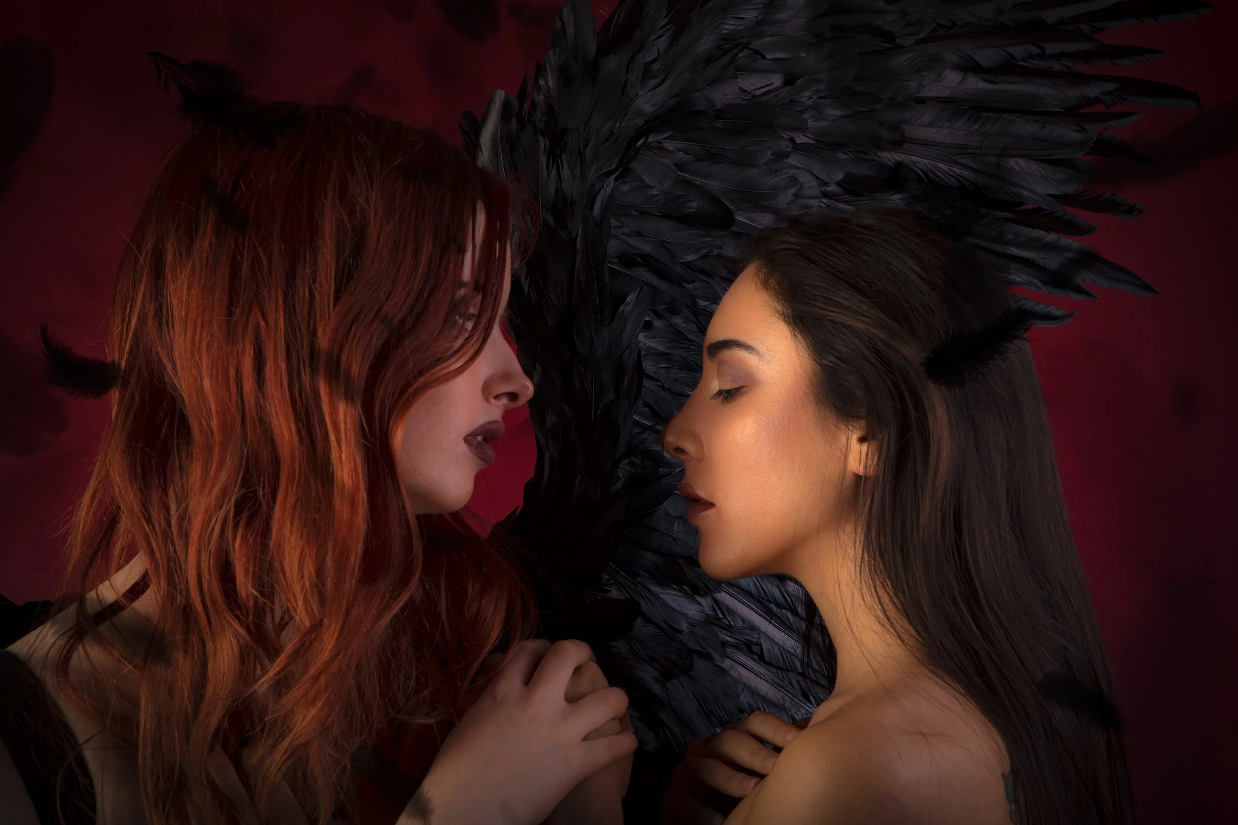 Two women face each other with their eyes closed, one with red hair and the other with dark hair, surrounded by black feathered wings against a dark red background.