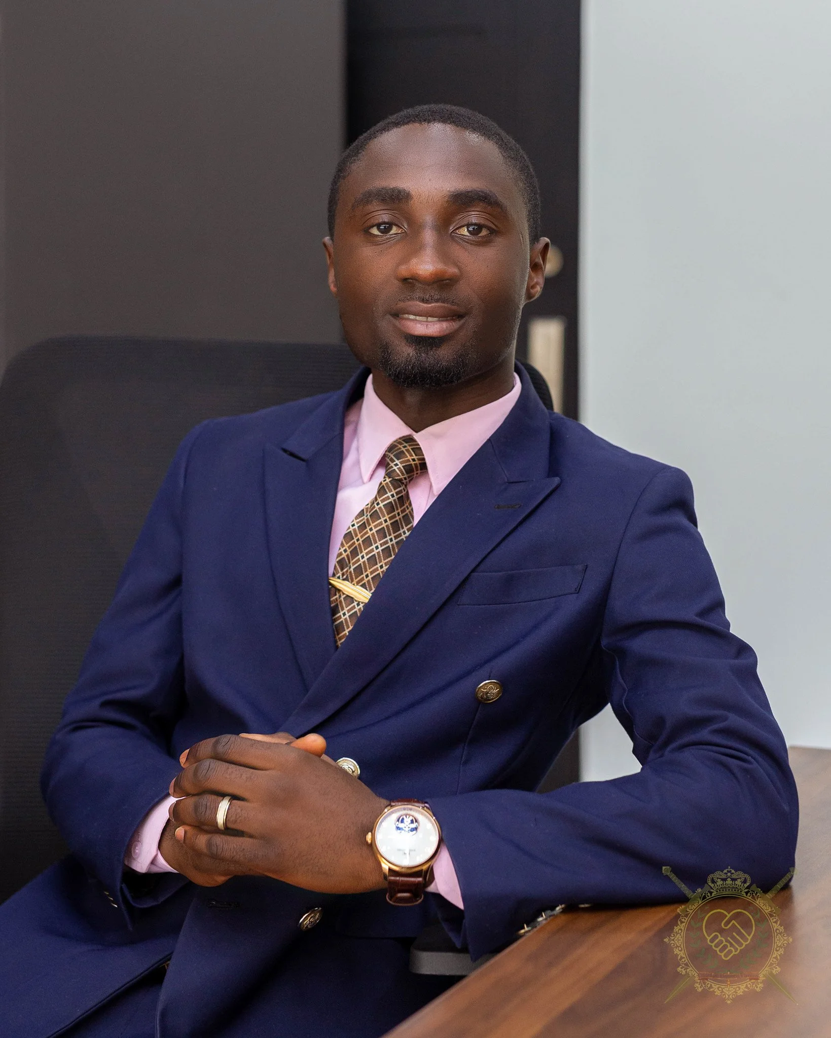 A young man in a dark blue suit with a pink shirt and patterned tie, sitting at a desk with hands clasped, wearing a watch and rings, in an office setting.