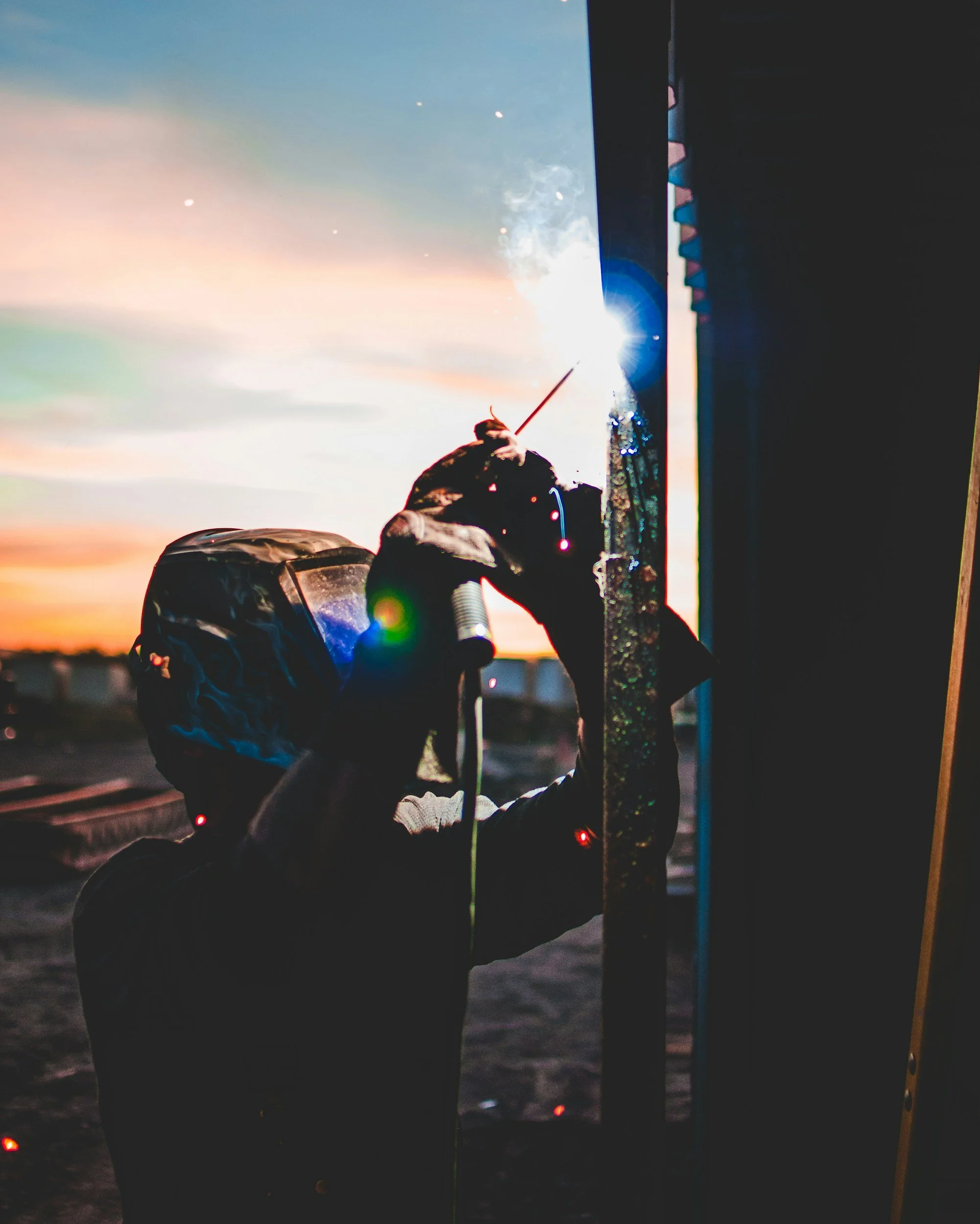 A welder working outdoors at sunset, wearing protective gear and welding metal with sparks flying.