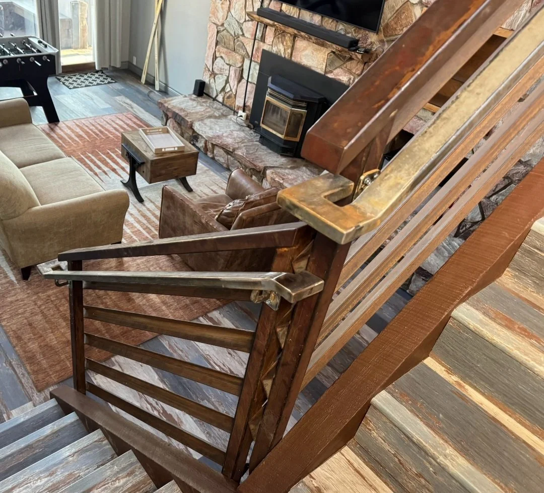View of a living room with a stone fireplace, a flat-screen TV mounted above, beige and brown furniture, a wooden coffee table, and wooden stairs with a custom metal handrail.