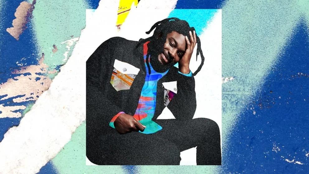 A man with dreadlocks and a beard, wearing a colorful patterned shirt and a black jacket, sits against a mixed blue and textured background, resting his head on his hand and smiling.