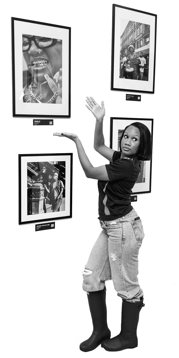 A woman in casual jeans and boots poses with her hand raised in front of framed black and white photographs displayed on a wall at an art gallery.