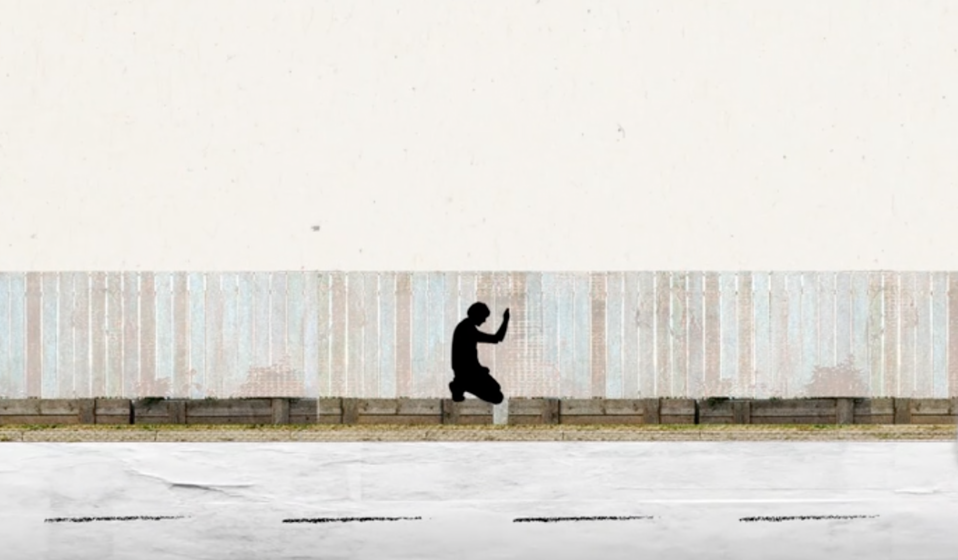 A person in silhouette kneeling on the sidewalk, raising one hand, with a large off-white wall in the background.