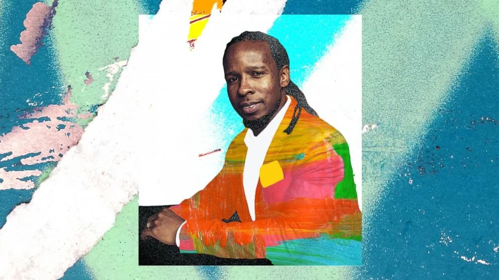A man with dark skin, long black hair in dreadlocks, wearing a colorful blazer with red, green, and yellow patches, and a white shirt, sitting against a backdrop of abstract watercolor textures in blue, green, pink, and white.