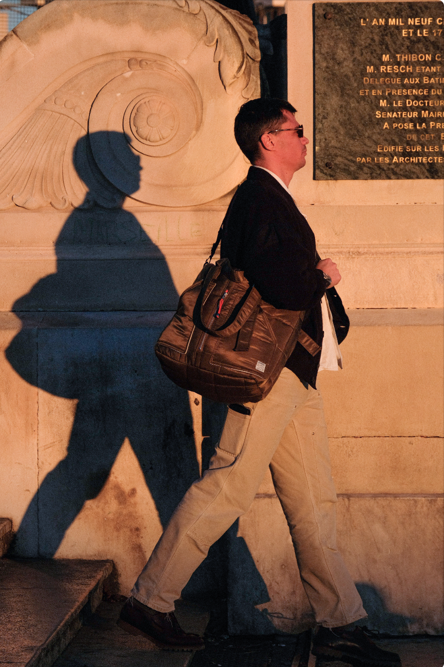 Un homme marche le long d'un mur, portant un sac à dos, portant des lunettes de soleil, avec une ombre projetée sur le mur derrière lui, sous une lumière orangée, dans une ville.