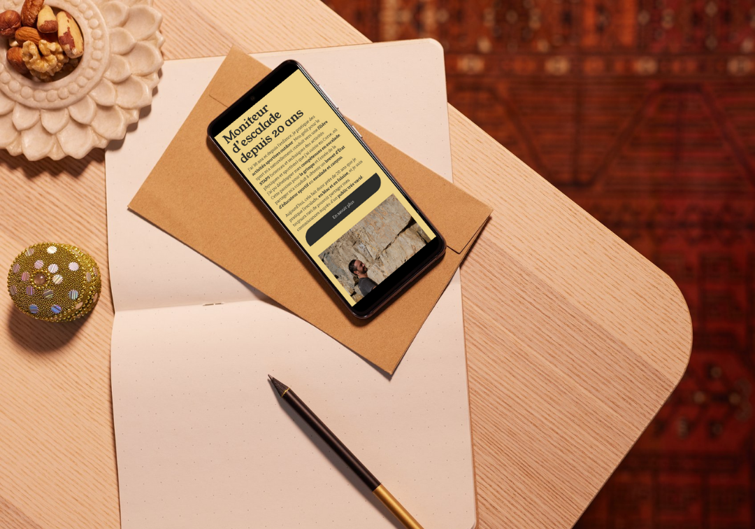 Une table en bois avec un téléphone, une enveloppe en papier craft, un carnet à pages pointillées, un stylo, un bol avec des noix et un fruit rouge décoré de paillettes.