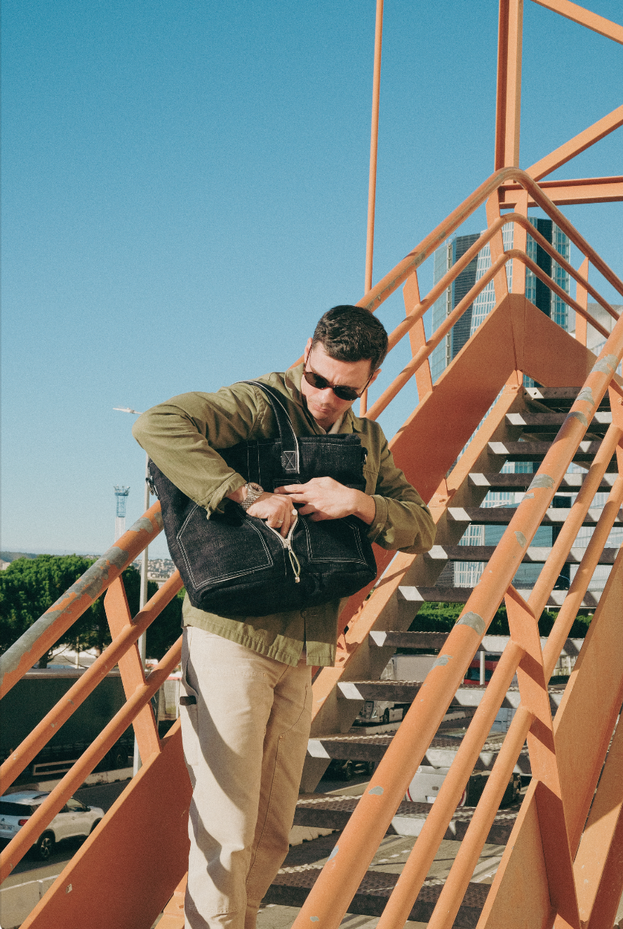 Un homme portant des lunettes de soleil, une veste verte et un pantalon beige, tient un sac à main noir en montant ou en descendant un escalier extérieur orange, avec un ciel bleu clair en arrière-plan.