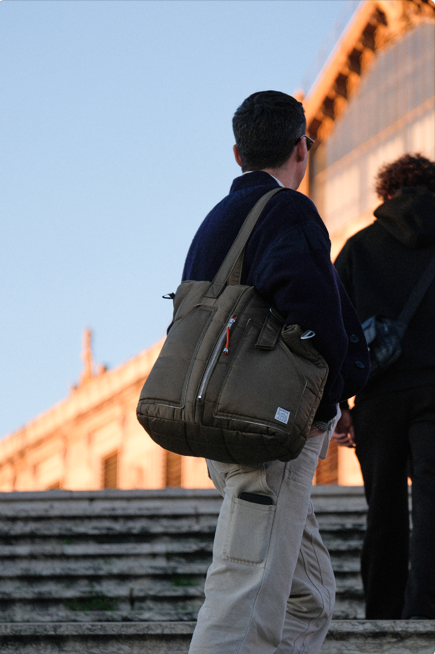 Un homme portant un sac à dos beige, vu de dos, debout devant un bâtiment ancien en pierre pendant le crépuscule.