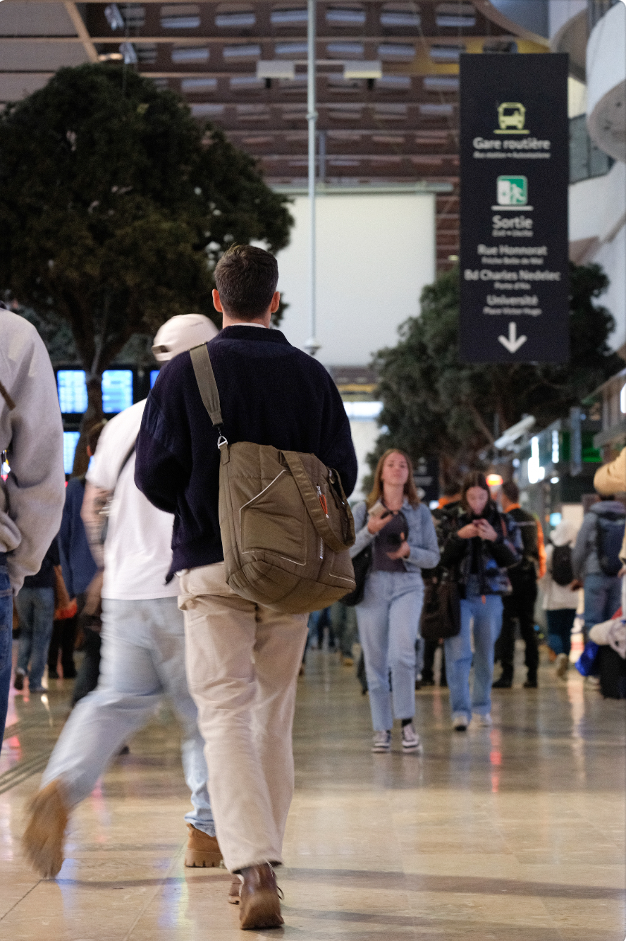 Personne marchant dans une gare avec d'autres personnes en arrière-plan, panneau d'informations en français à droite.
