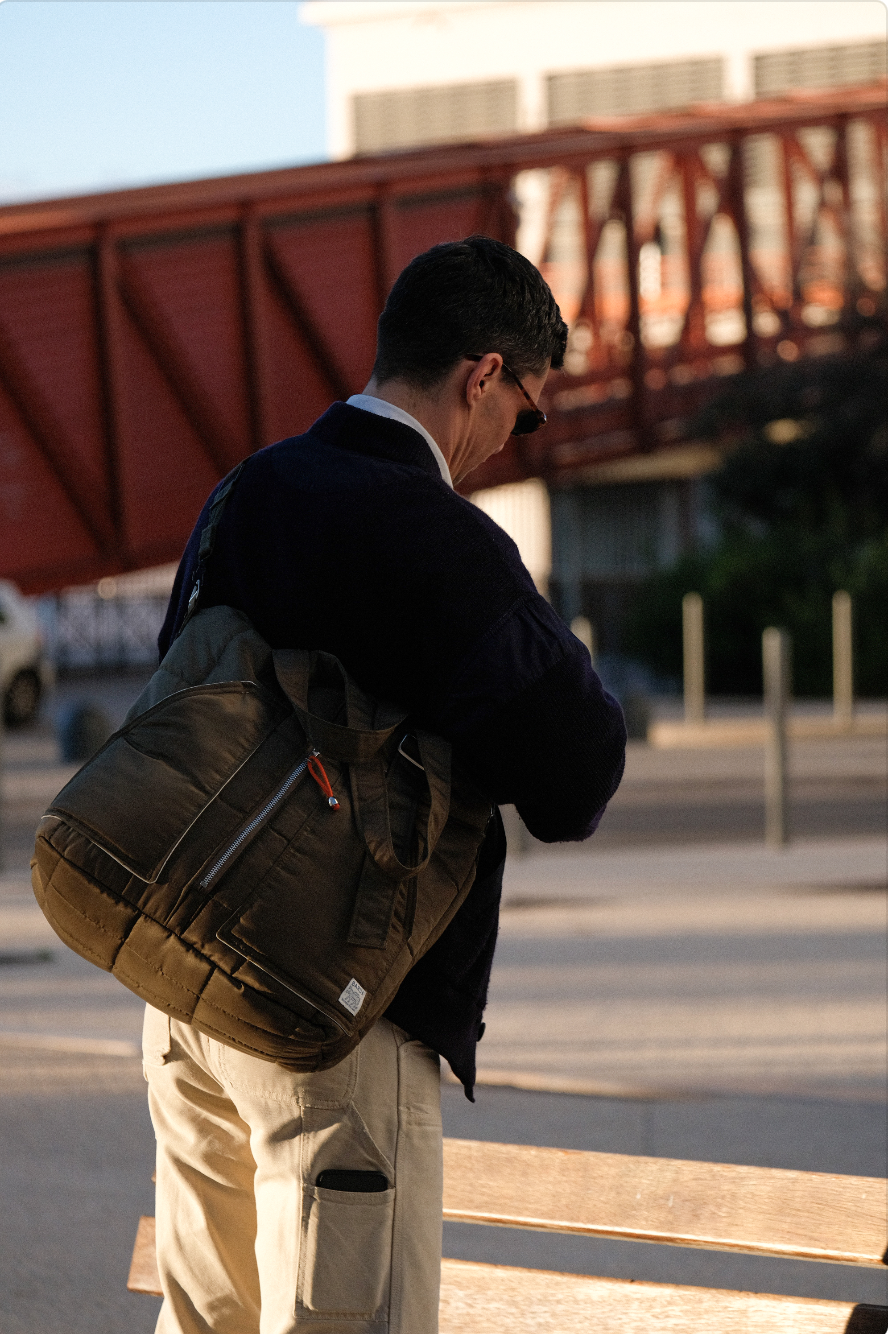 Un homme portant une veste noire, un sac à dos beige et des lunettes de soleil, se tient devant un banc en bois dans un environnement urbain au coucher du soleil.