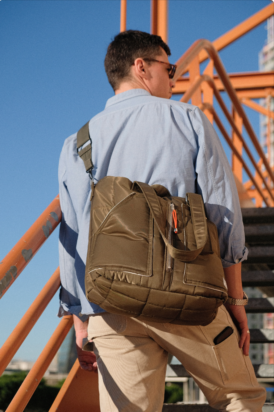 Jeune homme portant un sac à dos beige, en train de monter des escaliers extérieurs. Il porte des lunettes de soleil, une chemise bleu clair et un pantalon beige, avec un fond de ciel bleu et une structure en métal orange.