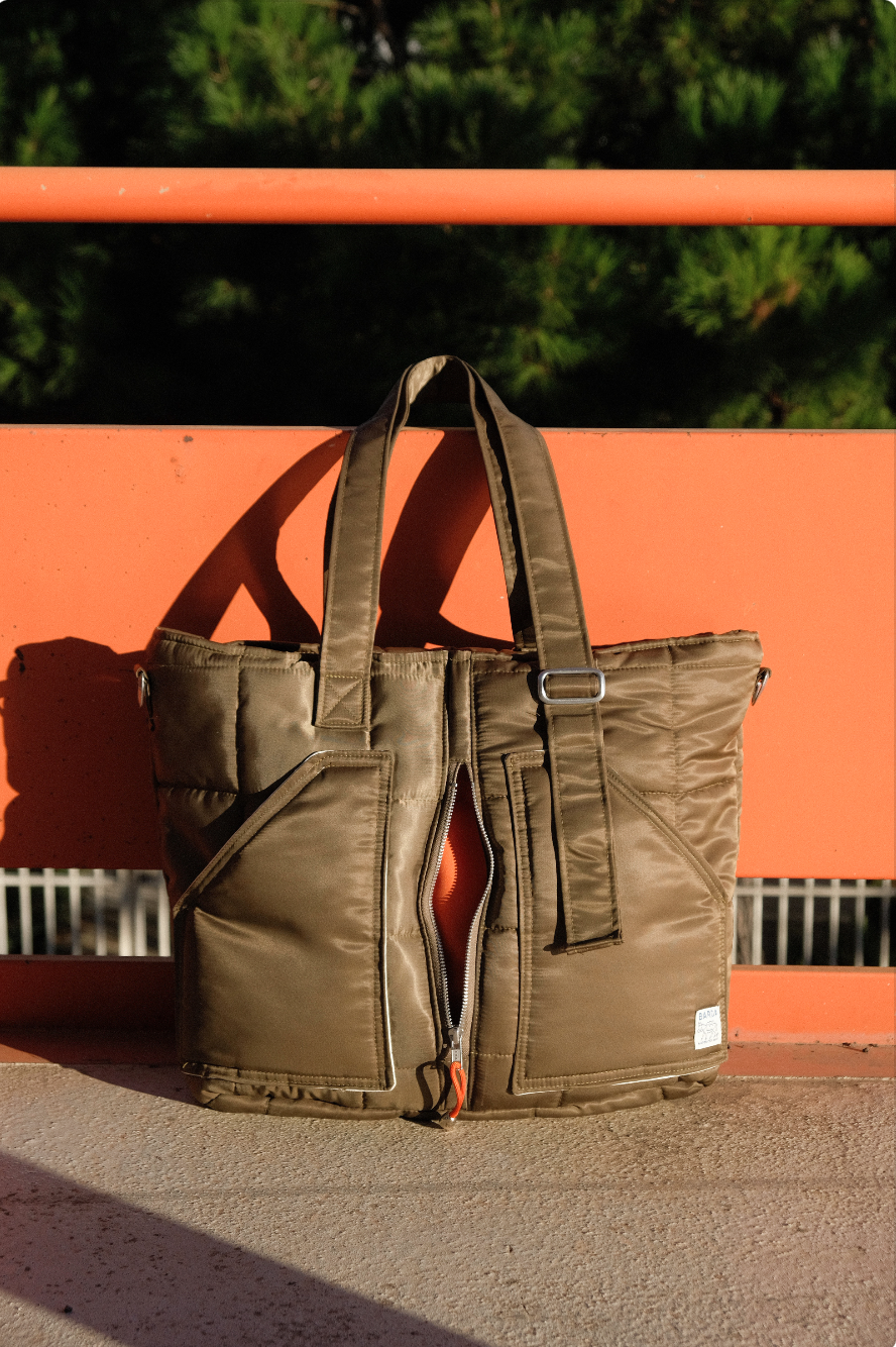 Sac à main en tissu matelassé de couleur kaki posé sur un banc en bois orange, avec des poches frontales, un zip et des poignées doubles, à l'extérieur.