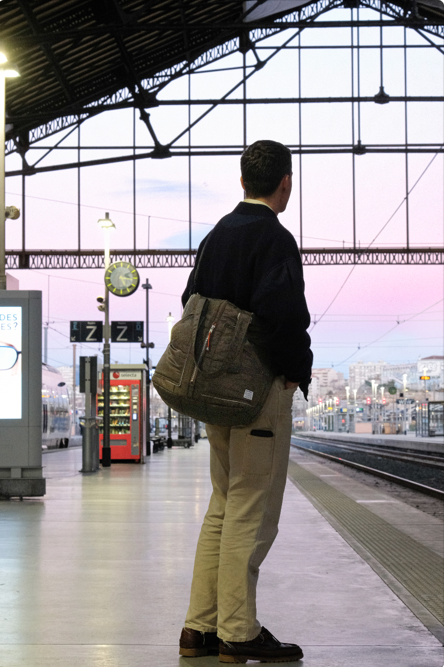 Un homme debout dans une gare, regardant au loin, portant un sac à dos, avec un ciel pastel au coucher du soleil visible à travers le toit en verre de la gare.