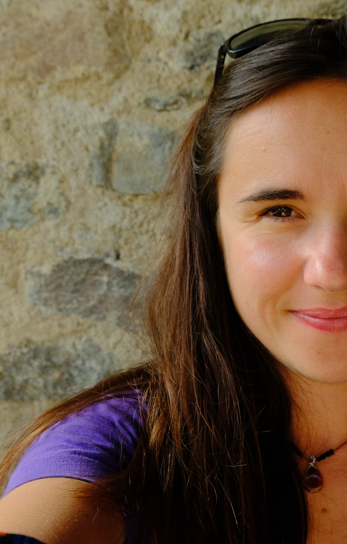 Rosalie une jeune femme femme souriante avec des cheveux longs, portant des lunettes sur la tête et un collier, devant un mur en pierre.