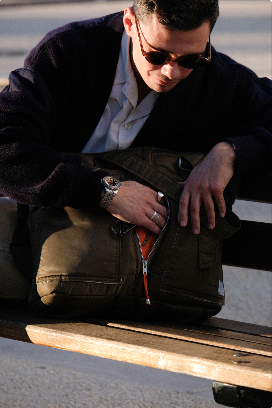 Homme portant des lunettes de soleil, debout à côté d'un skateboard, en train de ranger un sac à dos noir sur un banc.