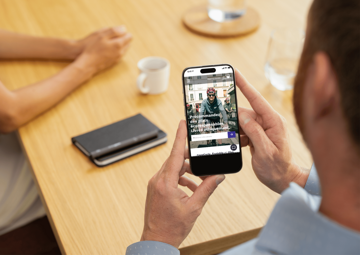 Une personne tient un smartphone affichant une page web sur un vélo, avec du texte en français. La scène se déroule à une table en bois avec une tasse blanche, deux livres en cuir et un verre d'eau.
