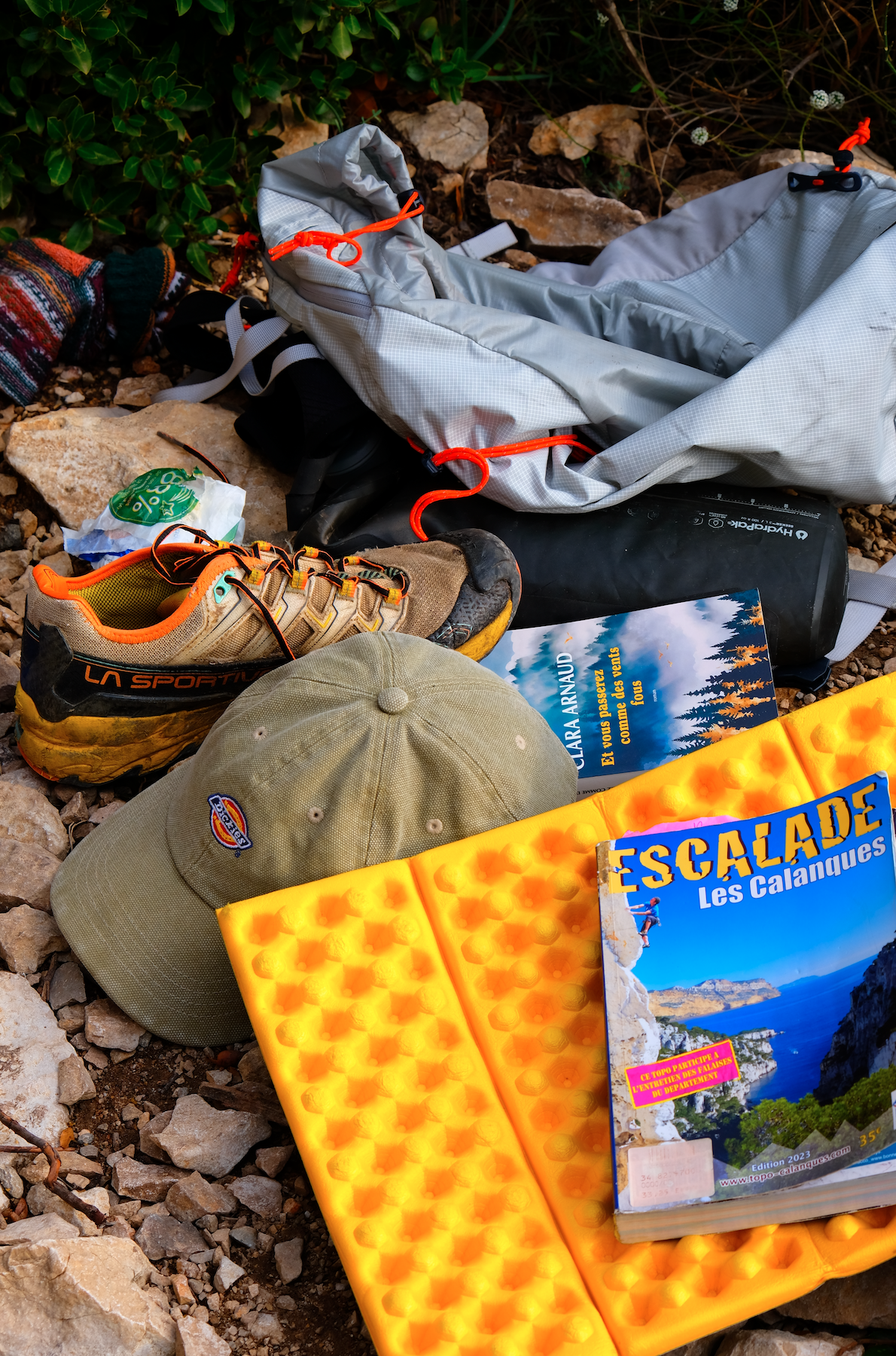 Matériel de randonnée sur le sol, comprenant une chaussure de marche, un chapeau, une casquette, un livre sur l'escalade, une alèse, une bouteille d'eau, un sac à dos, et des accessoires de camping. Il y a aussi un environnement naturel avec des pier