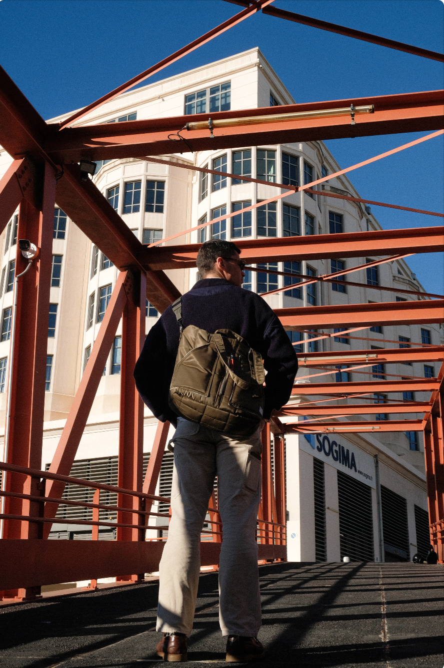 Un homme portant un sac à dos regarde vers un bâtiment moderne avec une façade blanche et de grandes fenêtres, sous un ciel bleu clair.