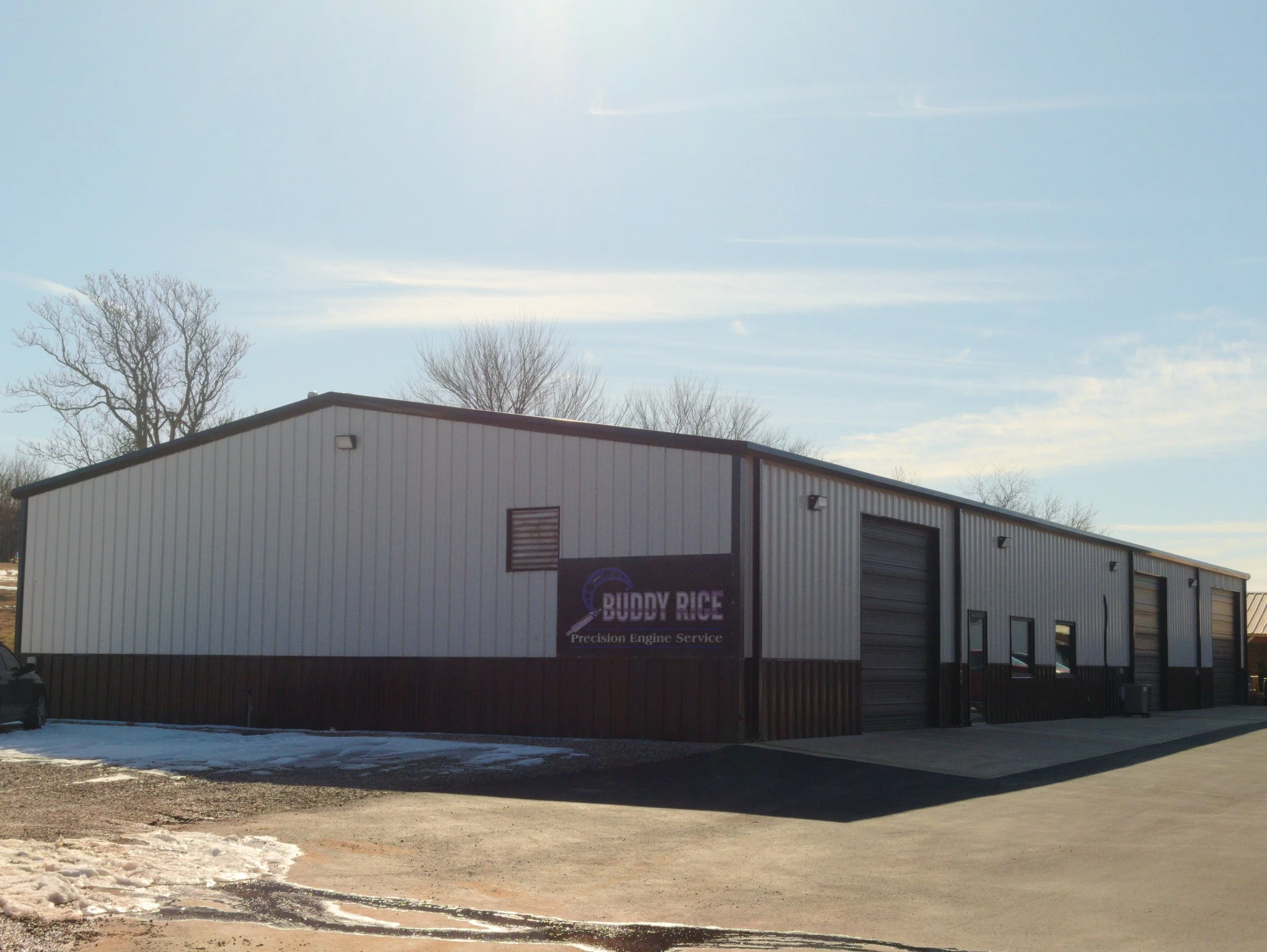 A large industrial building with metal siding, labeled 'Buddy Rice Precision Engine Service' on a sign on the front. There are several garage doors, some with windows, and the ground is partially covered with snow. The sky is clear with some clouds and leafless trees can be seen in the background.