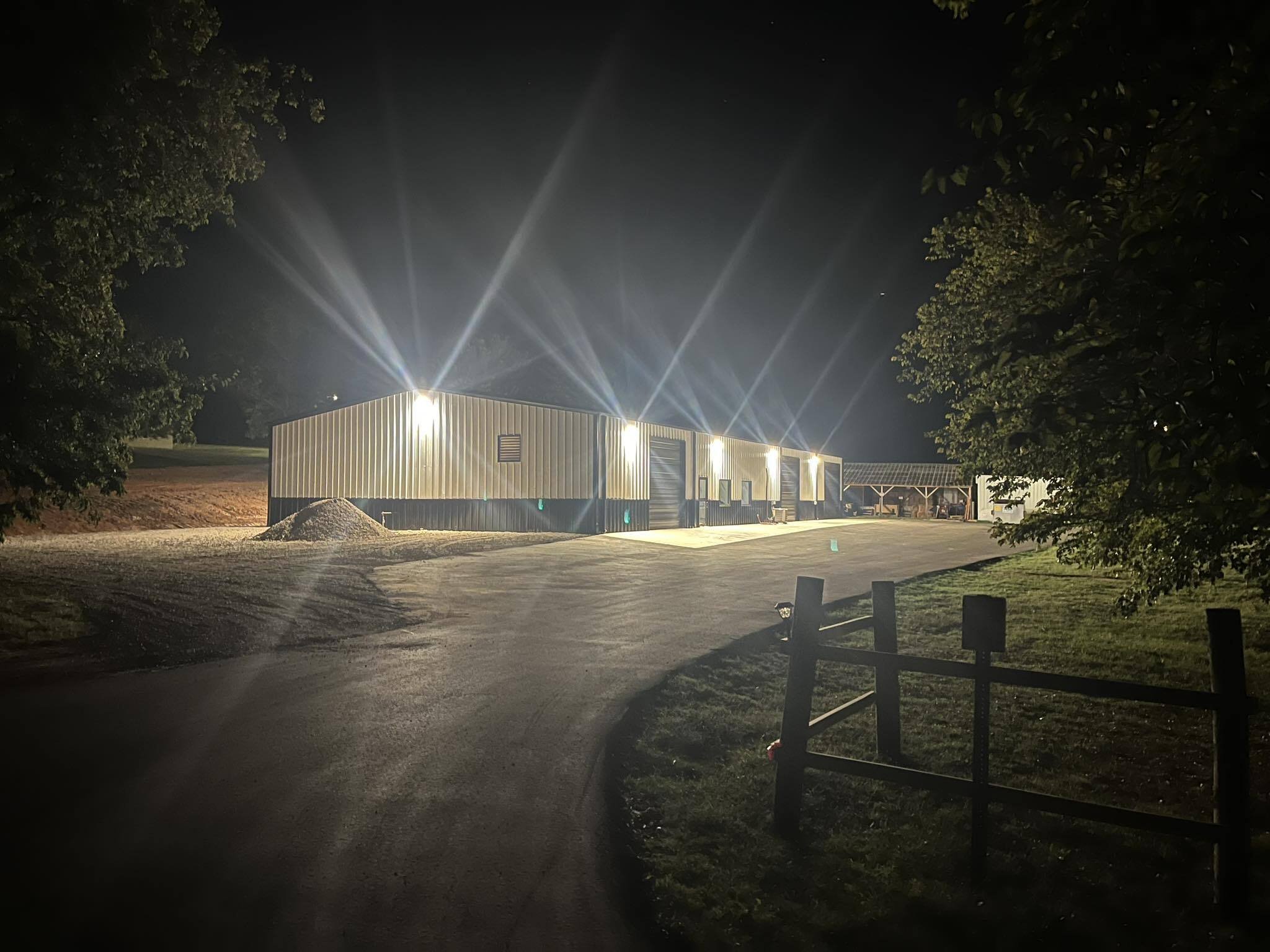 Nighttime scene of a large metal building with bright exterior lights creating starburst effects, surrounded by a gravel driveway, trees, and a small wooden fence.