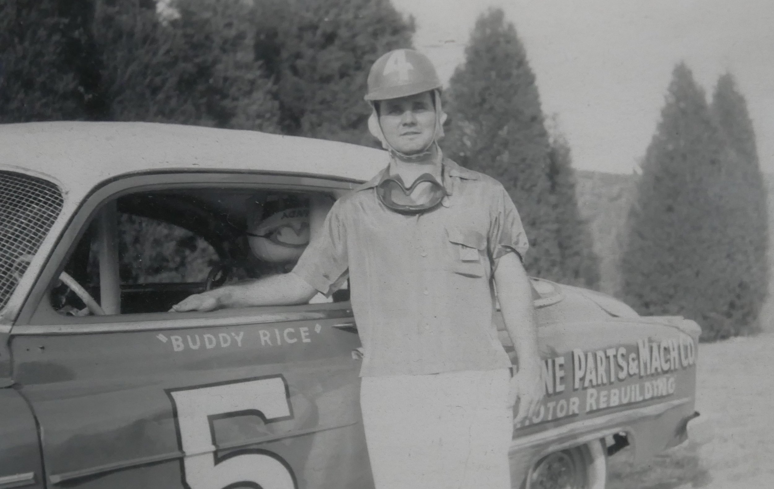 A young man in racing gear with a helmet and goggles stands next to a race car marked with the number 5 and the name 'Buddy Rice' on the side. The car has additional text indicating it is a motor rebuilding company. The background shows trees and an outdoor setting.