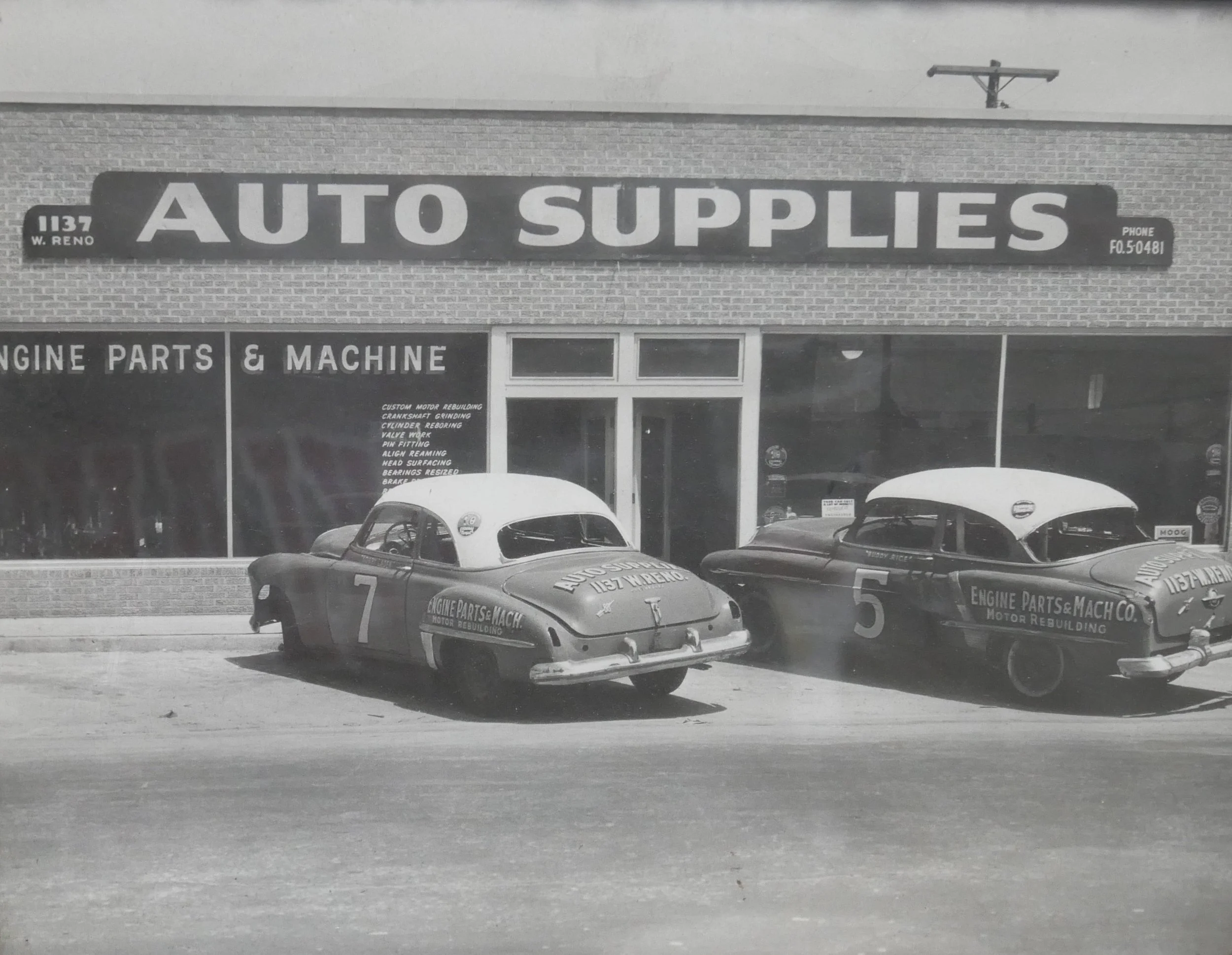 Black and white photo of two vintage race cars parked in front of an auto supplies shop with a sign reading 'Auto Supplies.' The cars have numbers 7 and 5 and are marked with 'Engine Parts & Mach.' The shop has large glass windows and a brick exterior.
