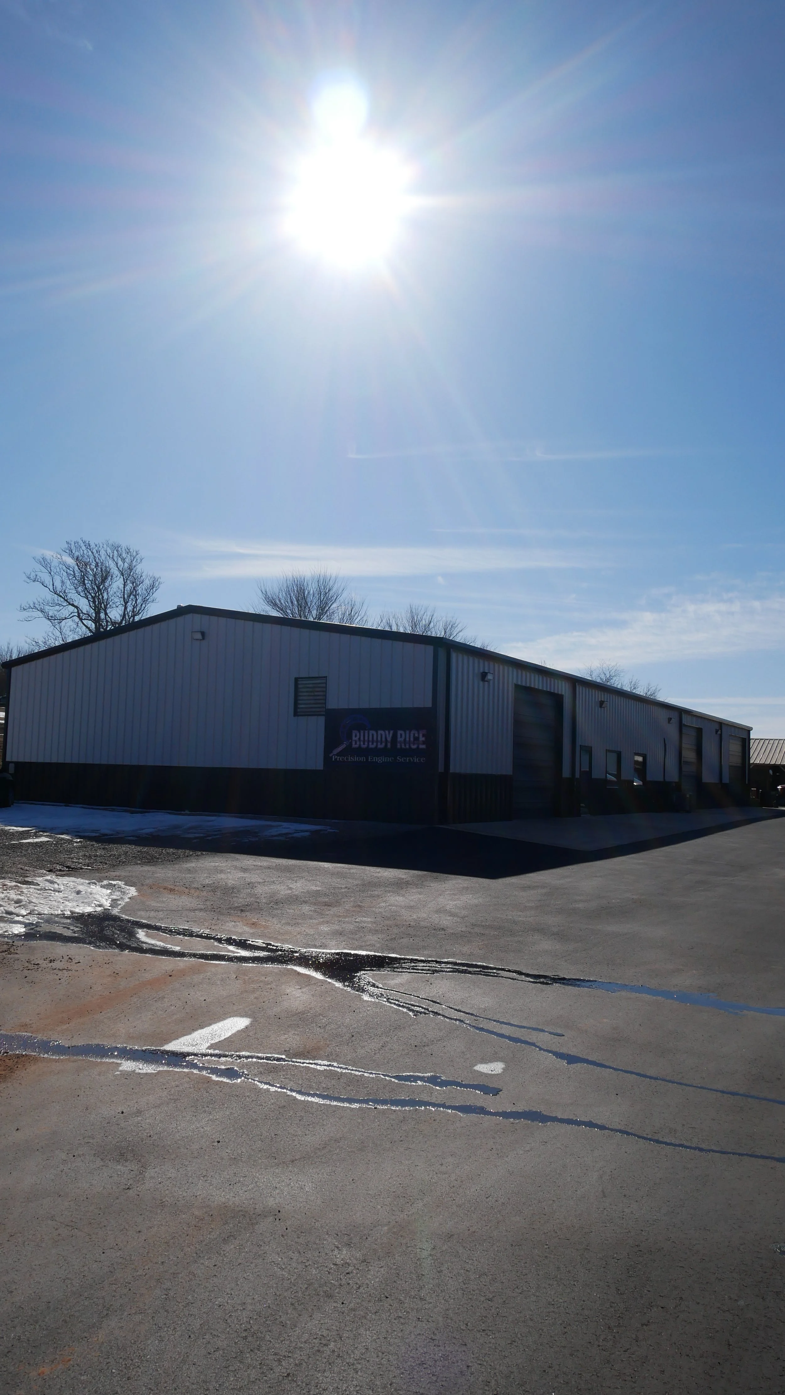 A large industrial building with a sign that reads 'Buddy Rice Precision Engine Service,' under a bright sun in a clear blue sky, with some snow on the ground and patches of water on the parking lot.