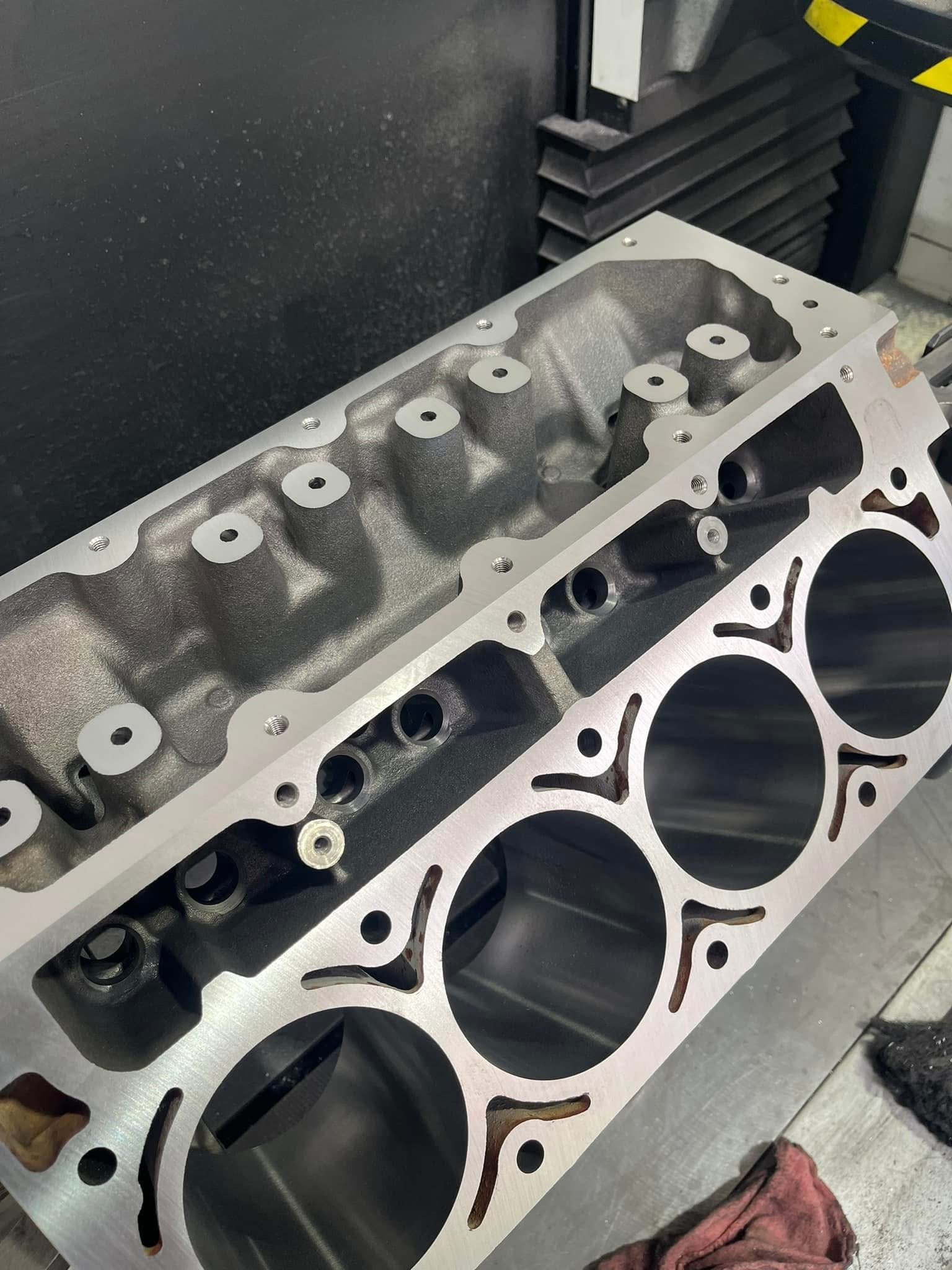 A partially assembled engine cylinder head made of metal, showing intake and exhaust ports, bolt holes, and combustion chambers on a workshop workbench.