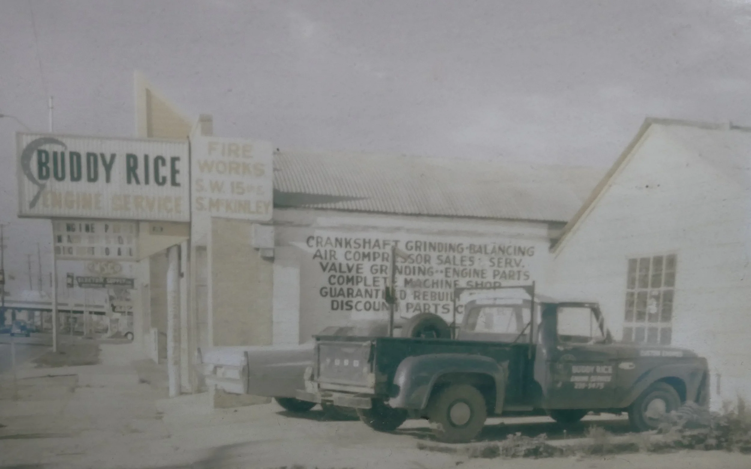 Black pickup truck parked in front of a building with signs for Buddy Rice Tire Service and fire works, with various service offerings listed on the wall.