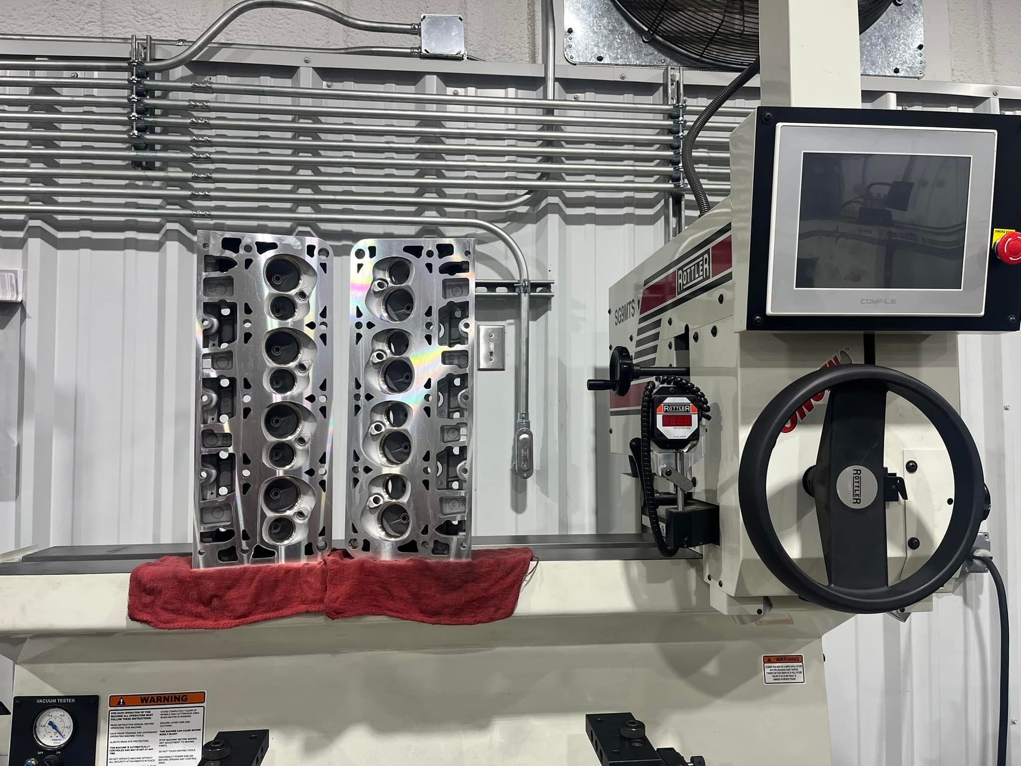 Two engine cylinder heads on a red towel in a machine shop, with a testing machine and monitor nearby.
