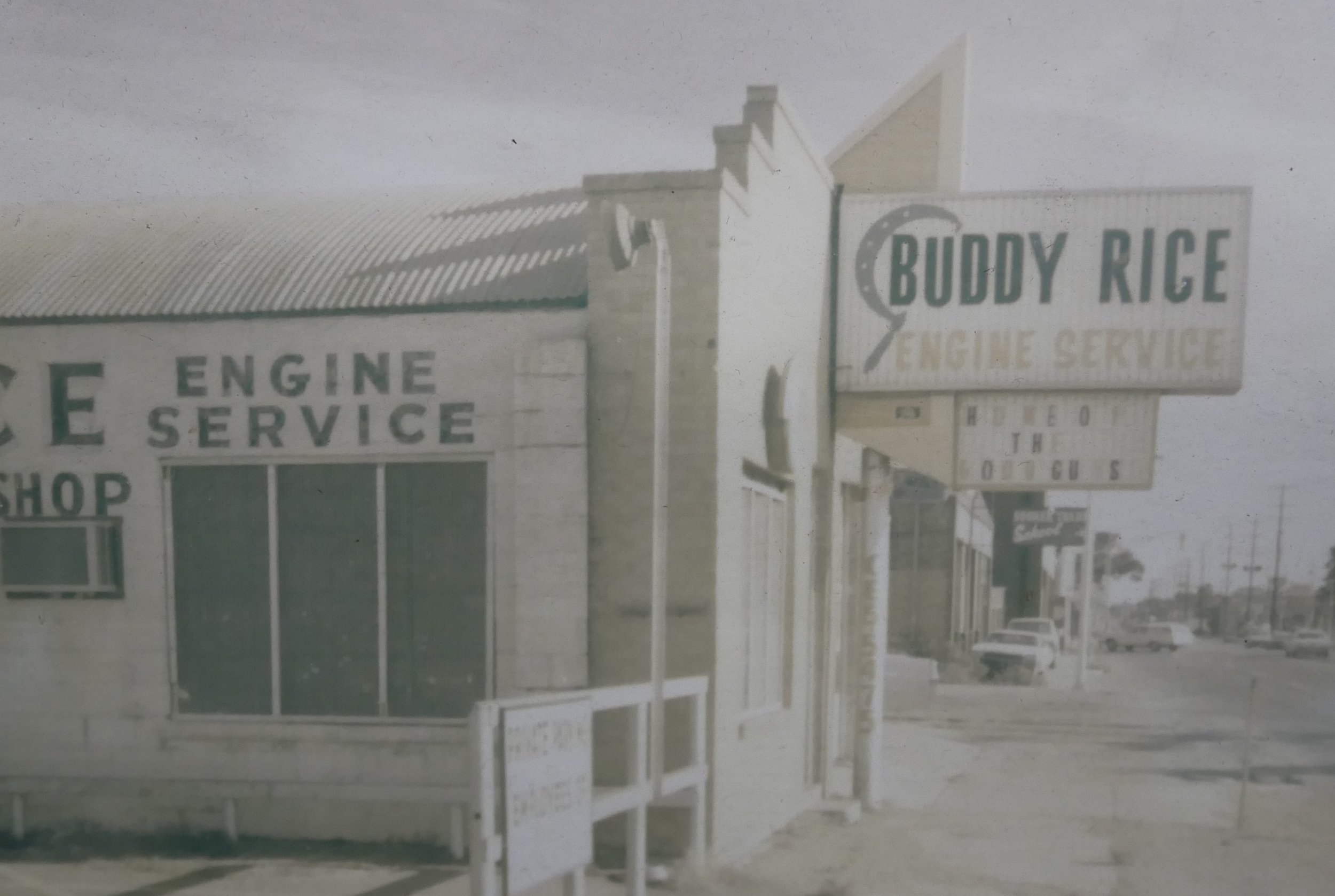 A vintage photo of Buddy Rige Engine Service with a large sign on the building and another sign on a pole advertising the same business. The building has a corrugated metal roof and a partially visible window. The street in front has parked cars and utility poles, suggesting a small town or suburban setting.