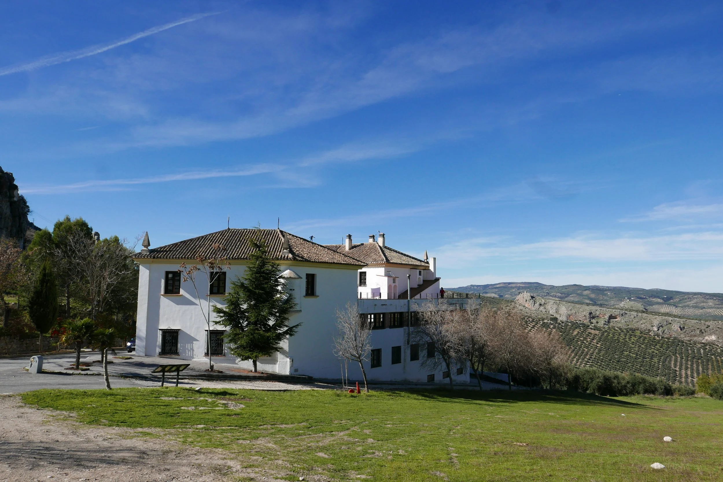 A white multi-story house with a tiled roof, set against a backdrop of rolling hills and clear blue sky. There are a few trees in the foreground, a small green lawn, and a pathway.