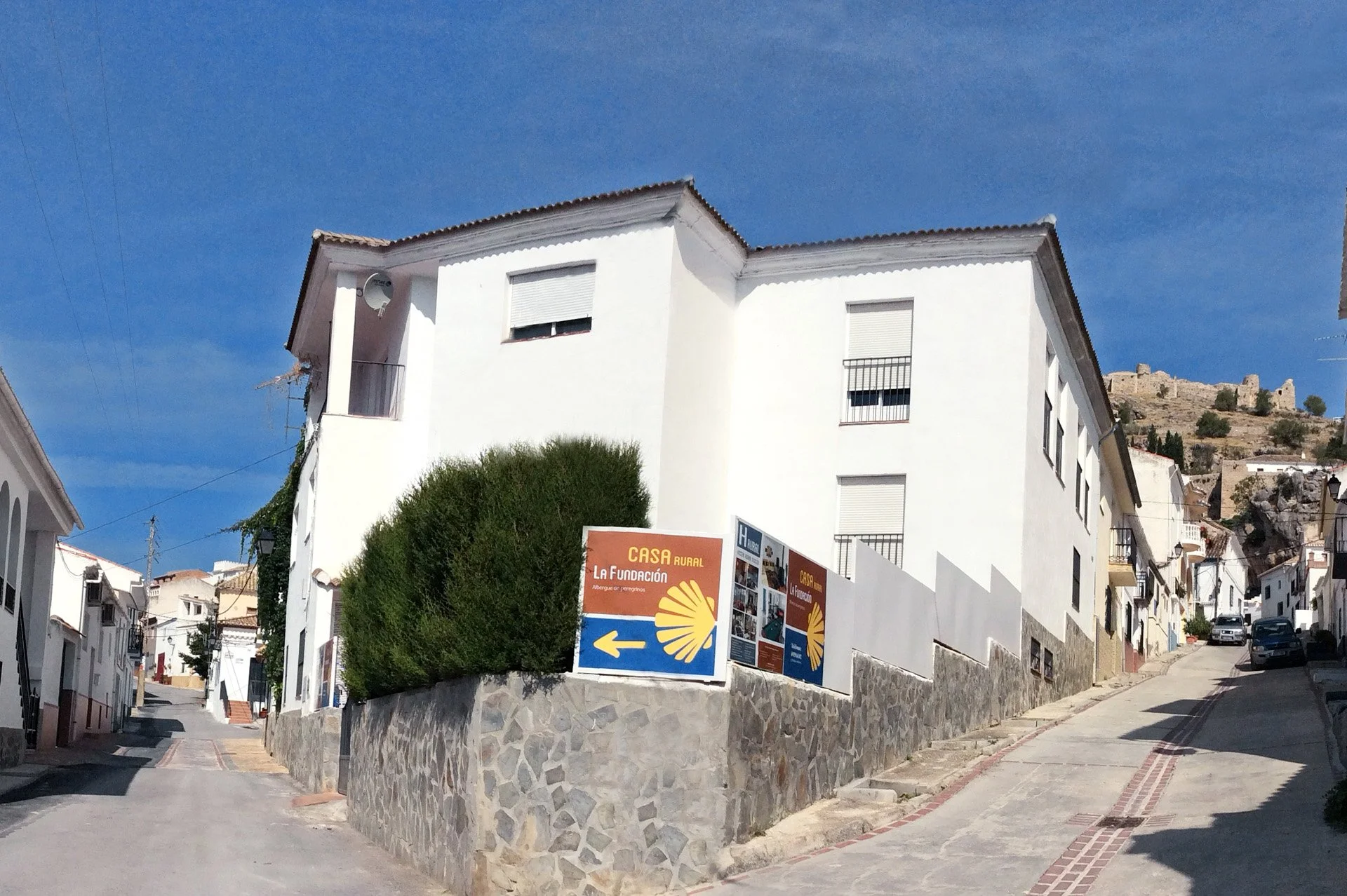 A white multi-story building with several windows, some with roller shutters, and a satellite dish on the balcony. There are signs on the wall in Spanish, and the street is narrow, winding uphill with a stone wall at the base. In the background, a hill with ruins and scattered trees under a clear blue sky.