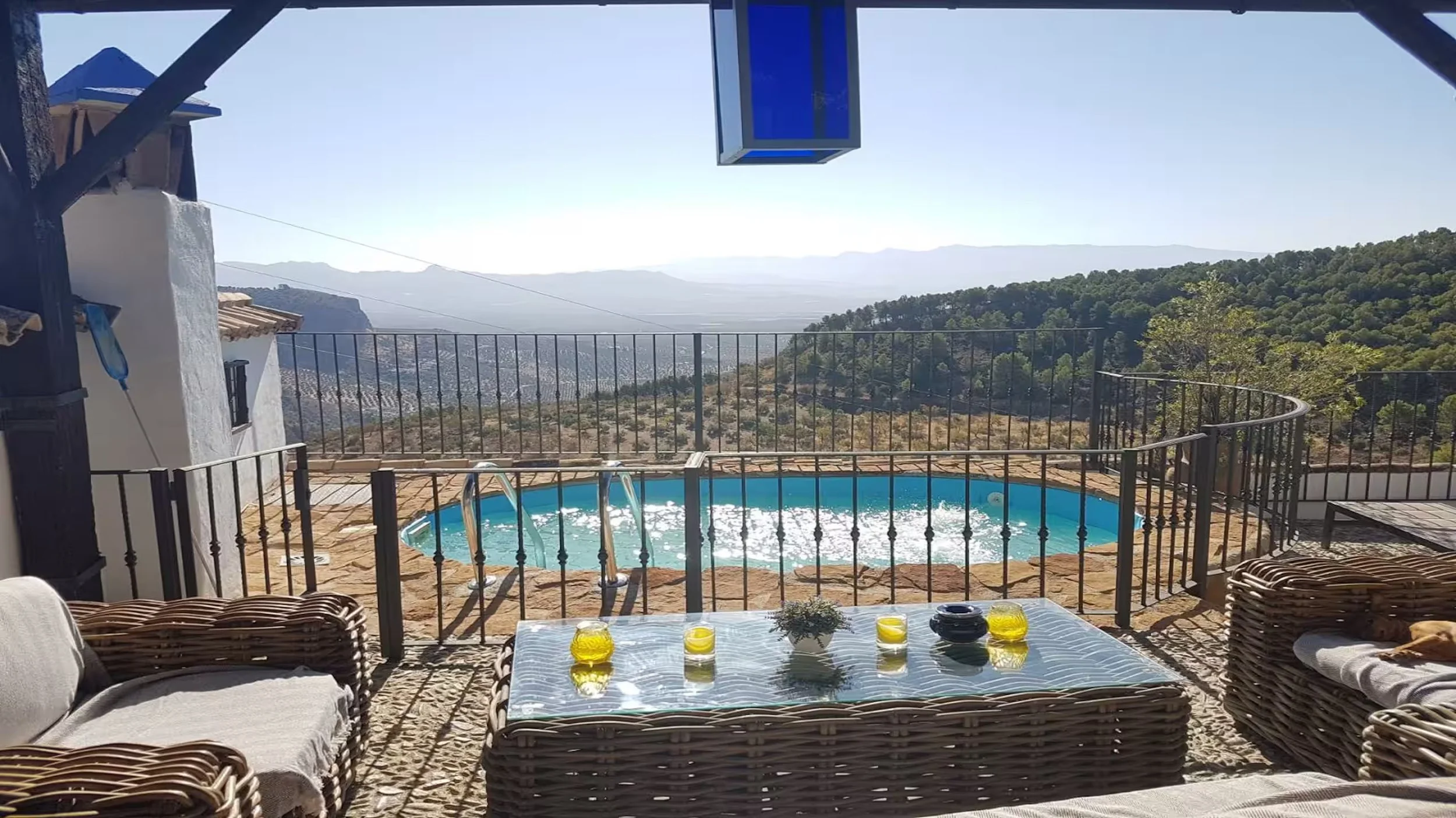 Patio with wicker furniture and a glass-topped table holding yellow candles, overlooking a fenced hot tub and scenic mountain landscape in the distance.