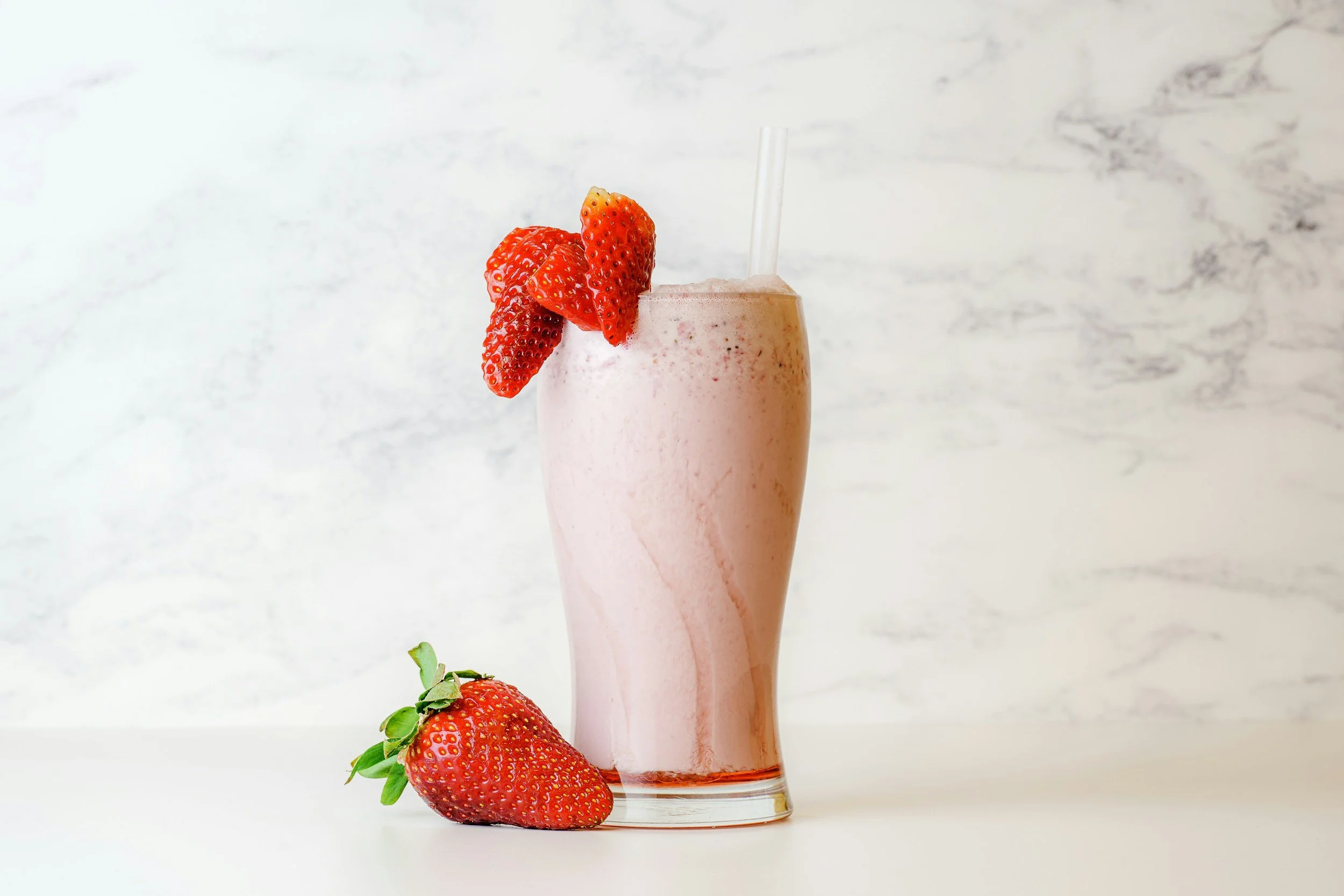 A strawberry smoothie in a tall glass garnished with sliced strawberries and a straw, with a fresh strawberry placed on the white surface beside the glass, against a white marble background.