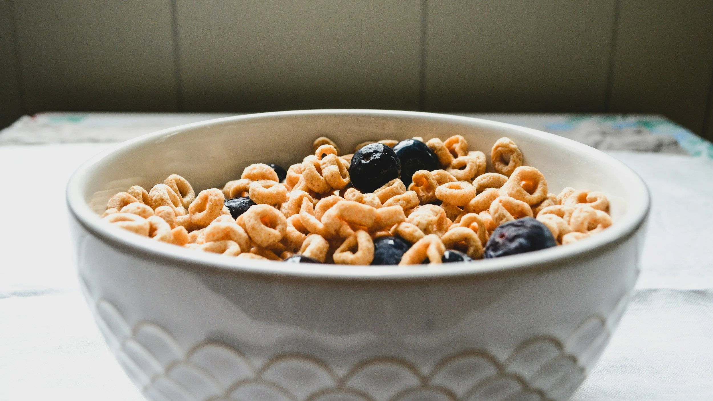 A white bowl filled with Cheerios cereal and blueberries on a white surface.