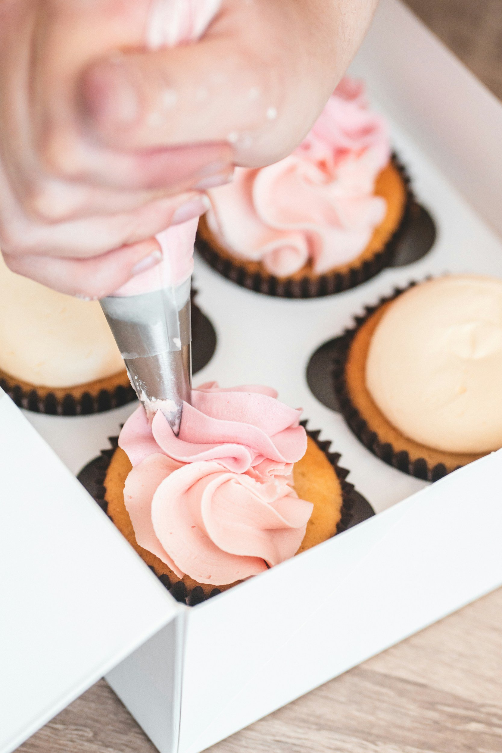 Person piping pink frosting onto cupcakes in a white box.