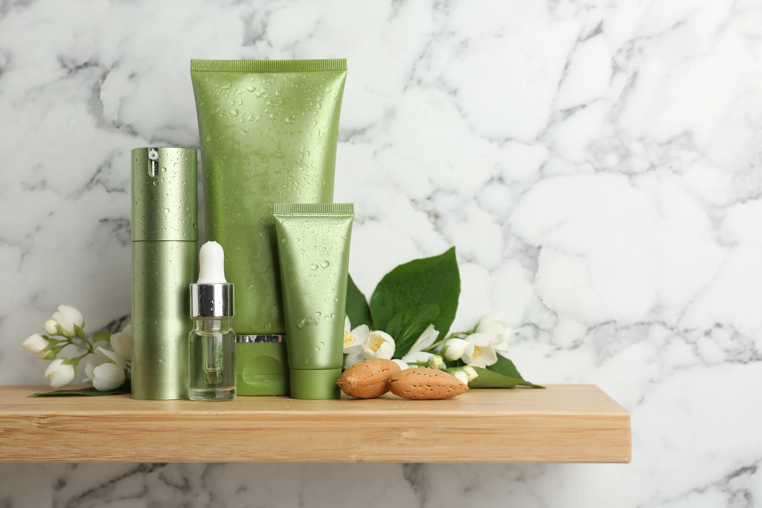 Collection of green skincare products, a small glass dropper bottle, almonds, and white flowers on a wooden shelf against a marble wall.