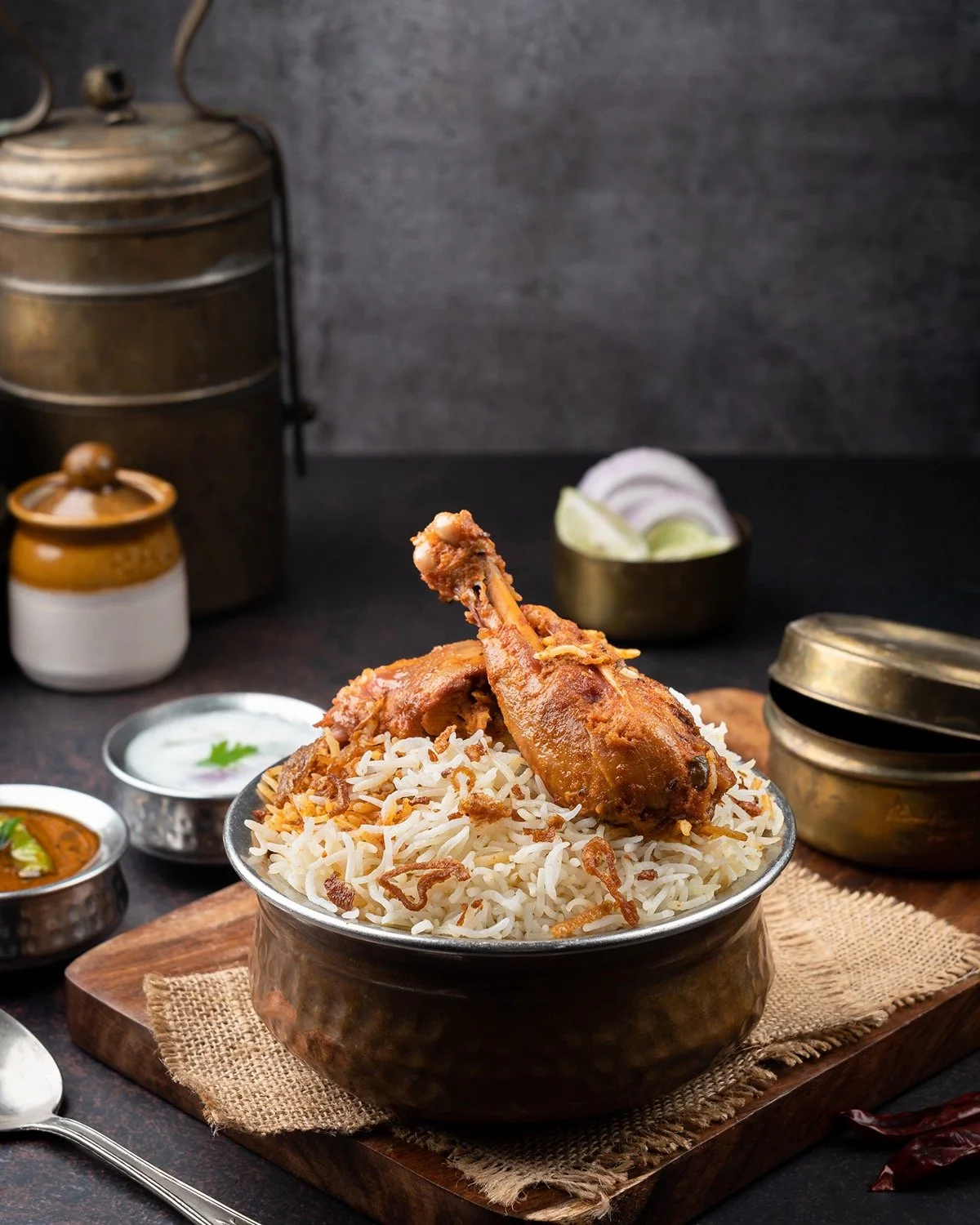A bowl of cooked rice with a cooked chicken leg on top, garnished with fried onions, on a wooden board, with small bowls of curry and spices around it.