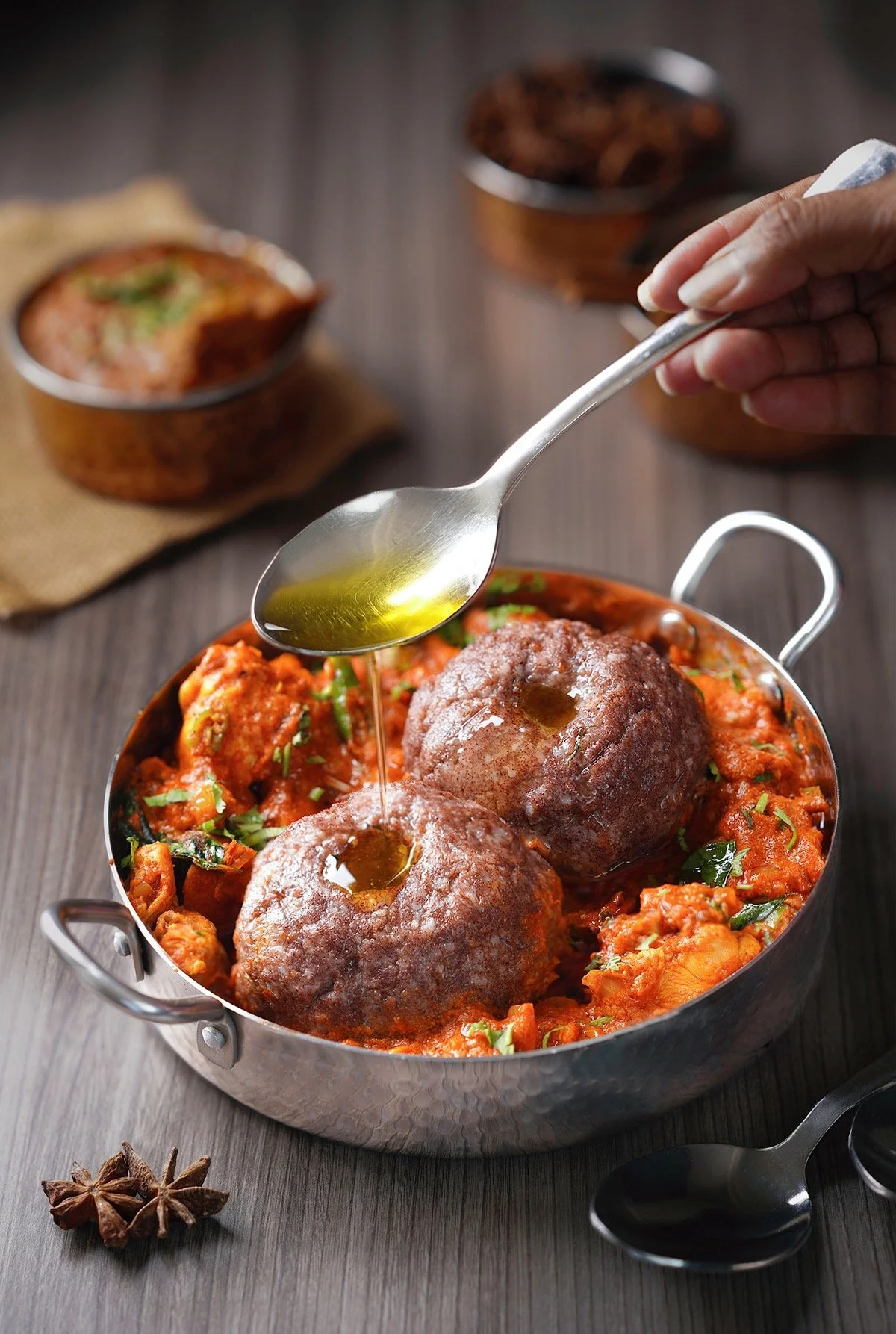 A metal dish of Indian-style meatballs in spicy tomato sauce, topped with a drizzle of oil, with star anise and additional bowls of food in the background.