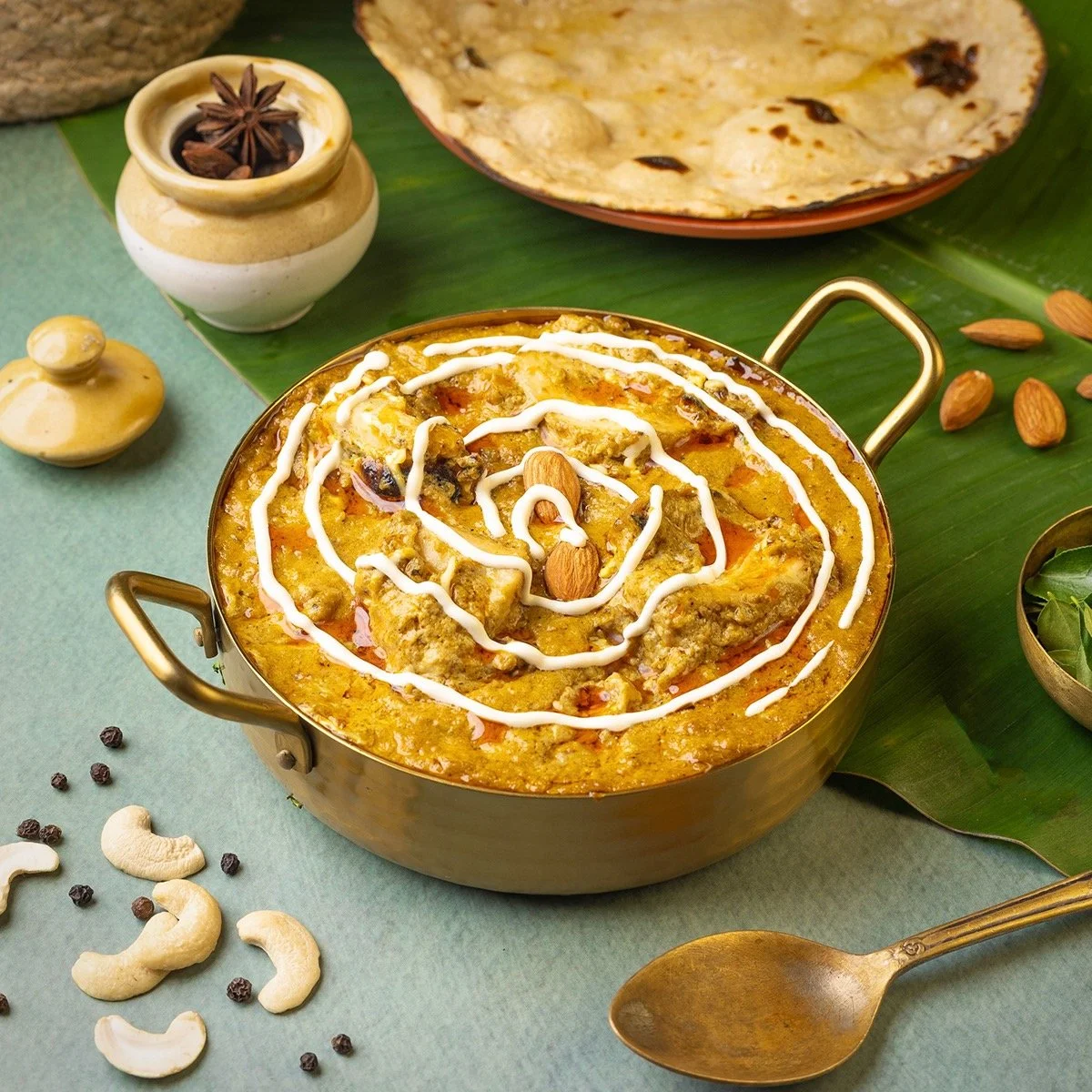 A bowl of Indian curry garnished with white sauce and almonds, surrounded by spices, cashews, and a notepad of flatbread on a green table.