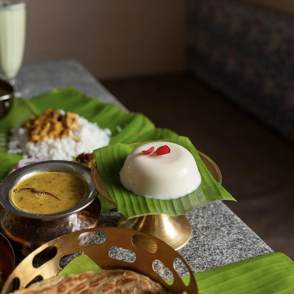 A traditional Indian dessert, a white pudding topped with rose petals, served on banana leaf. Surrounding items include a bowl of yellow custard and a plate with rice and curry, all presented on a granite countertop.