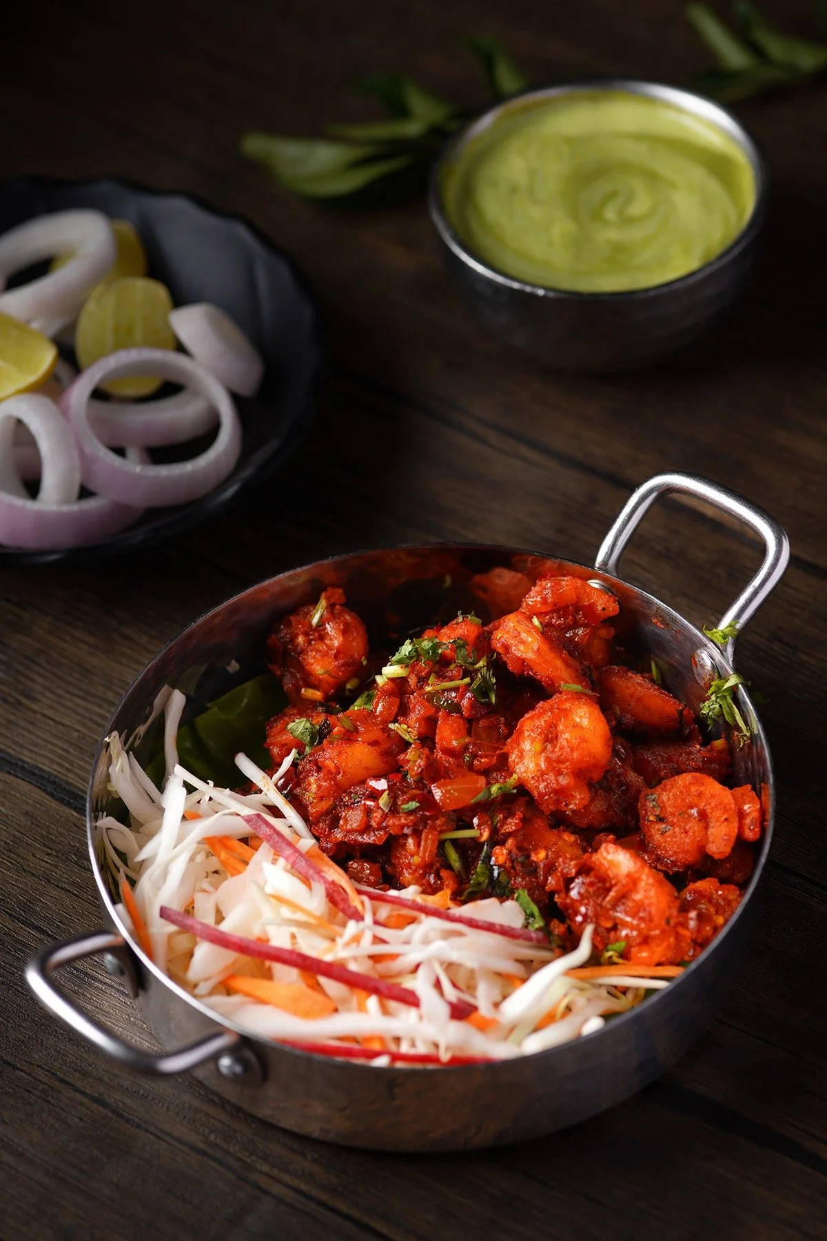 A plate of Indian curry with vegetables, a side of shredded cabbage and carrots, a bowl of green chutney, and sliced onions and lemon wedges on a black dish, all on a dark wooden table.