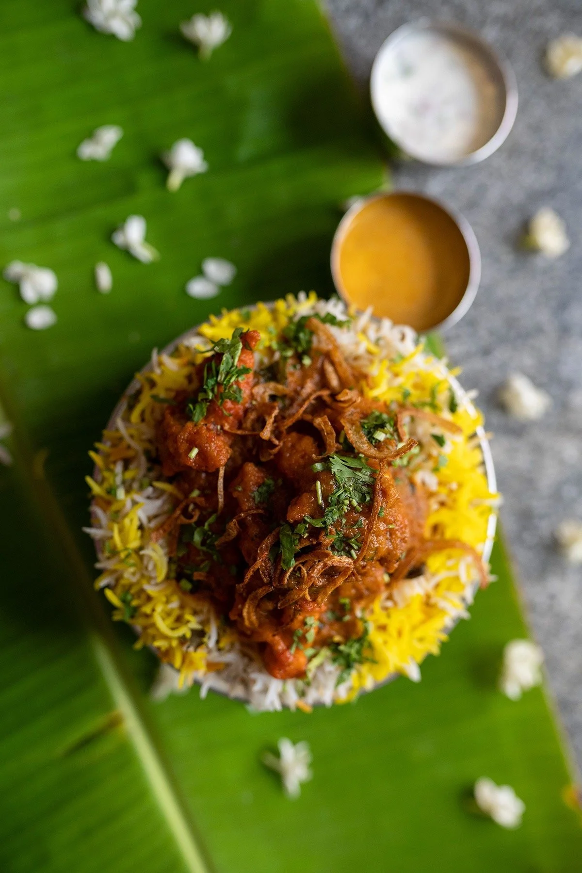 A plate of Indian food with rice, curry, and fried onions, garnished with herbs on a banana leaf, with small bowls of sauces and scattered flowers around.