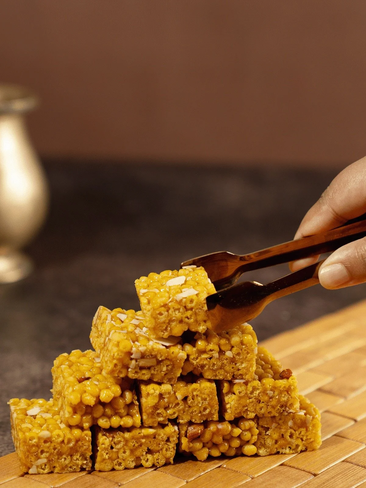 Stack of honey sesame bars on a wooden surface with a hand using tongs to pick one up.
