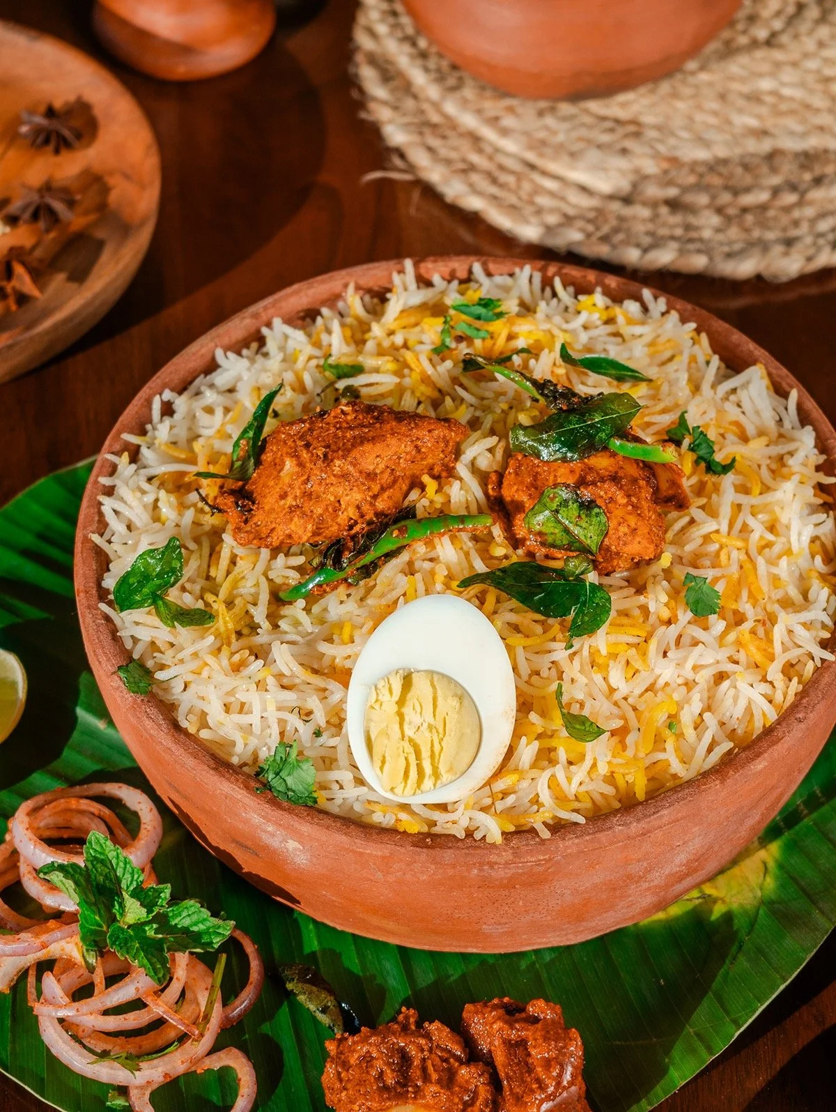A bowl of biryani rice with pieces of fried chicken, a halved boiled egg, and green herbs, served on a banana leaf.