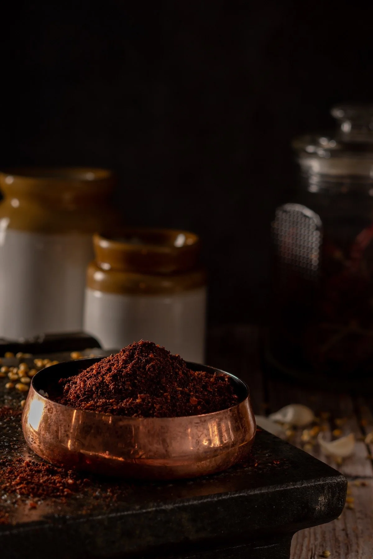 Copper bowl filled with ground coffee on a black cutting board, with various spices and ingredients faintly visible in the background.