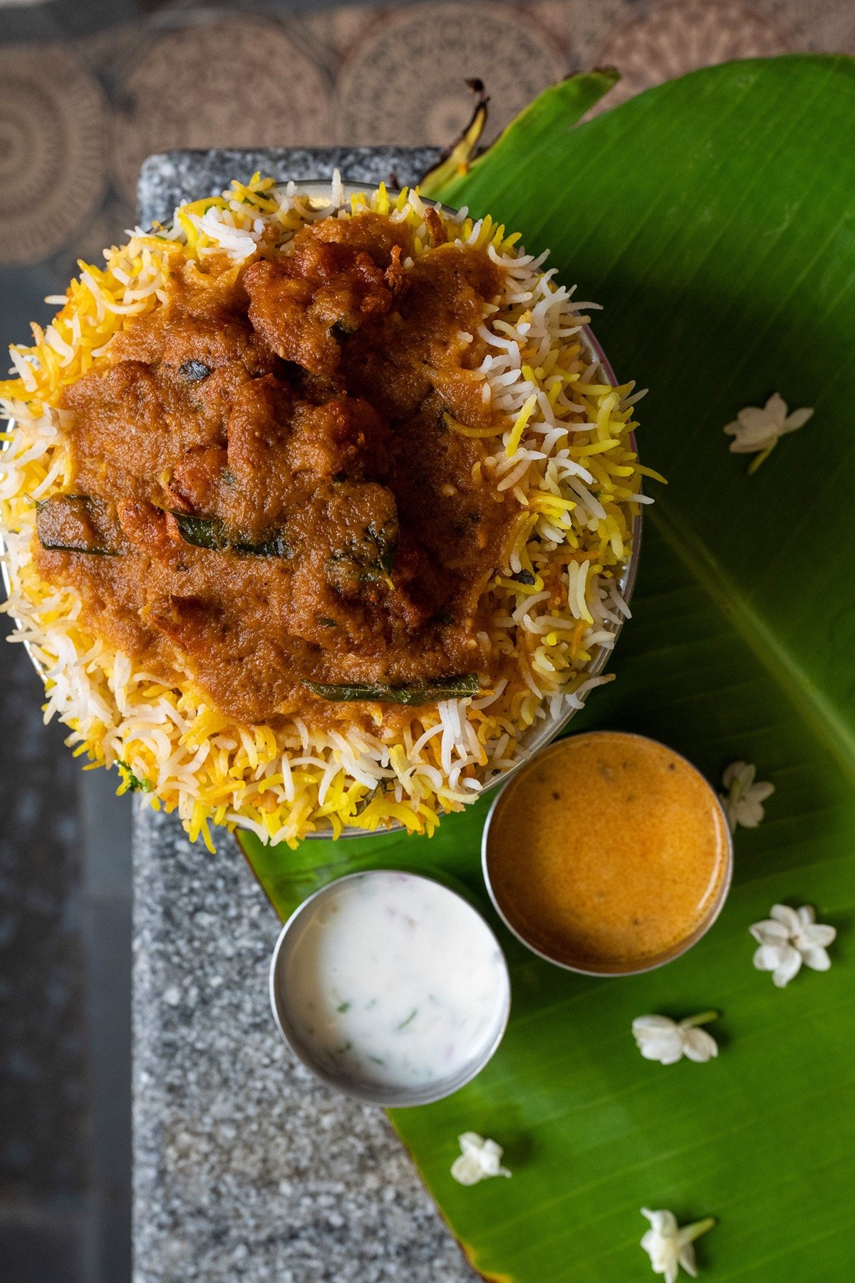A bowl of Indian rice biryani with chicken curry, served with white yogurt sauce and spicy orange sauce on a green leaf with small white flowers.