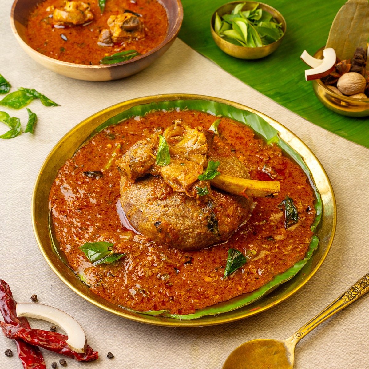 A bowl of Indian chicken curry with a potato and garnished with green curry leaves, served on a banana leaf with other bowls of spices and vegetables around.
